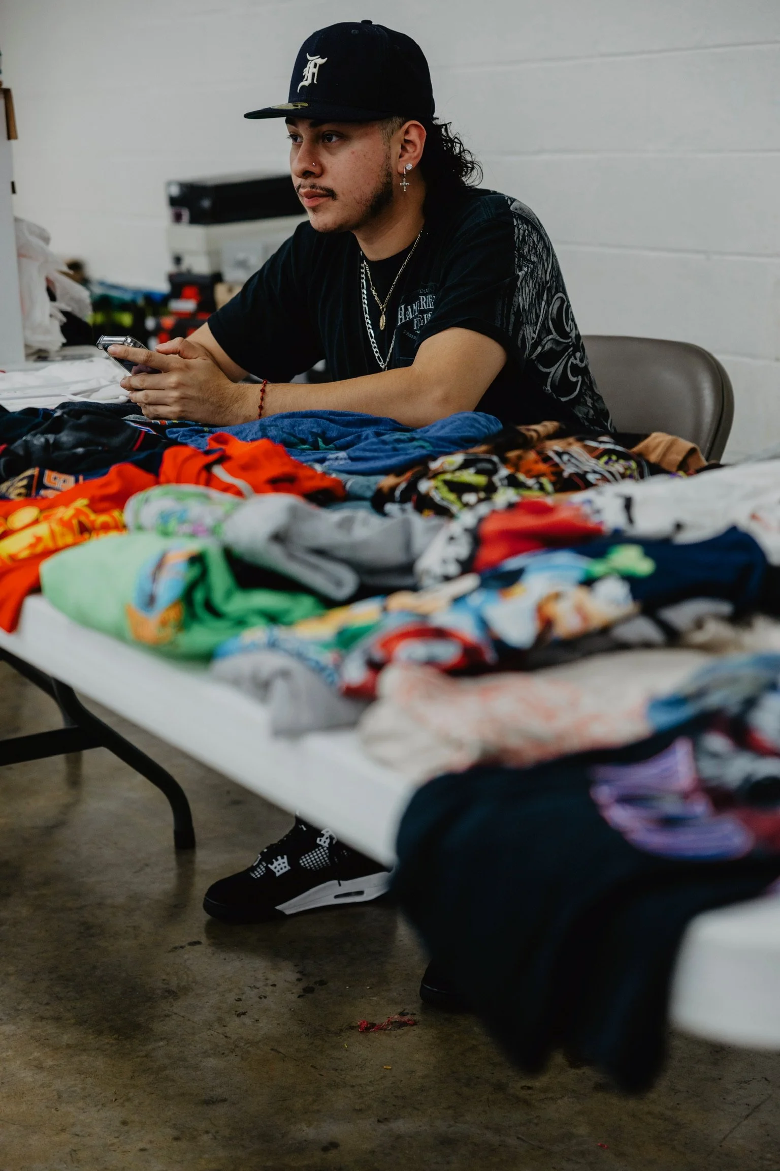 A man sitting at a table with colorful clothes and socks, wearing a black cap, black shirt, and jewelry, in an indoor setting.