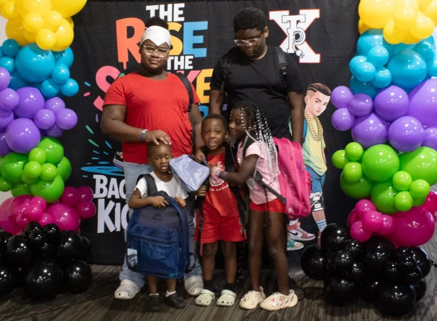 A group of two adults and three children posing in front of a colorful balloon wall at an event called 'The Rise of the Shadow' for back-to-school children.