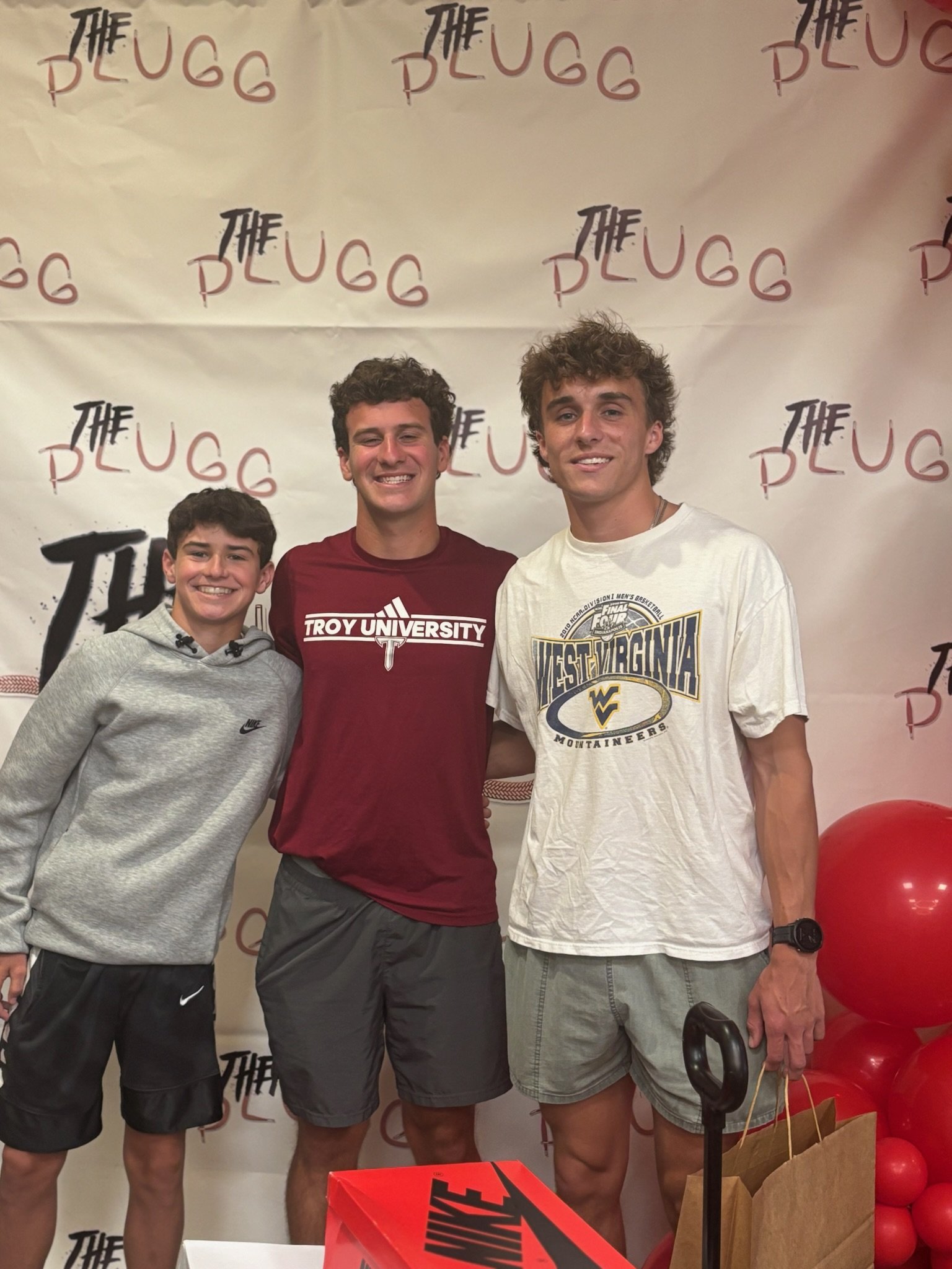 Three young men standing together indoors in front of a backdrop with 'The Puffg' written repeatedly, smiling at the camera. The man on the left is wearing a gray Nike hoodie, the middle man is wearing a maroon Troy University T-shirt, and the man on