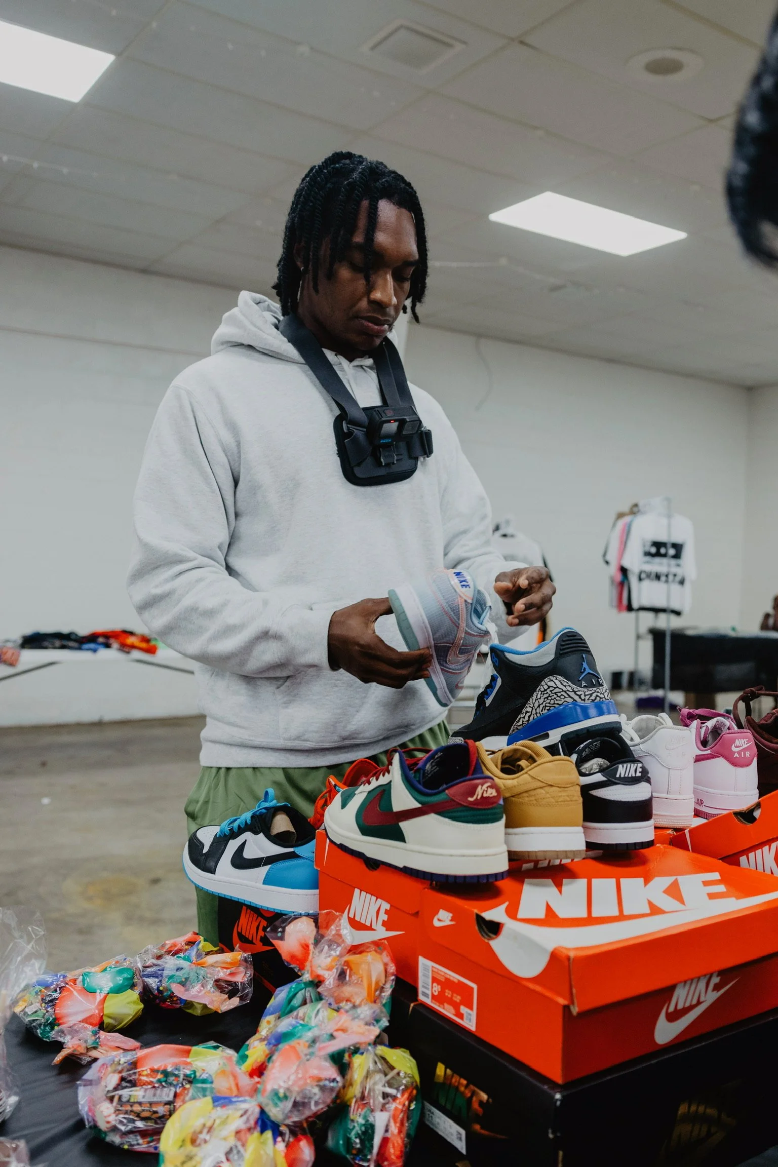 A man standing behind a table filled with colorful sneakers and candy, holding a white sneaker, in a room with white walls and a white ceiling.