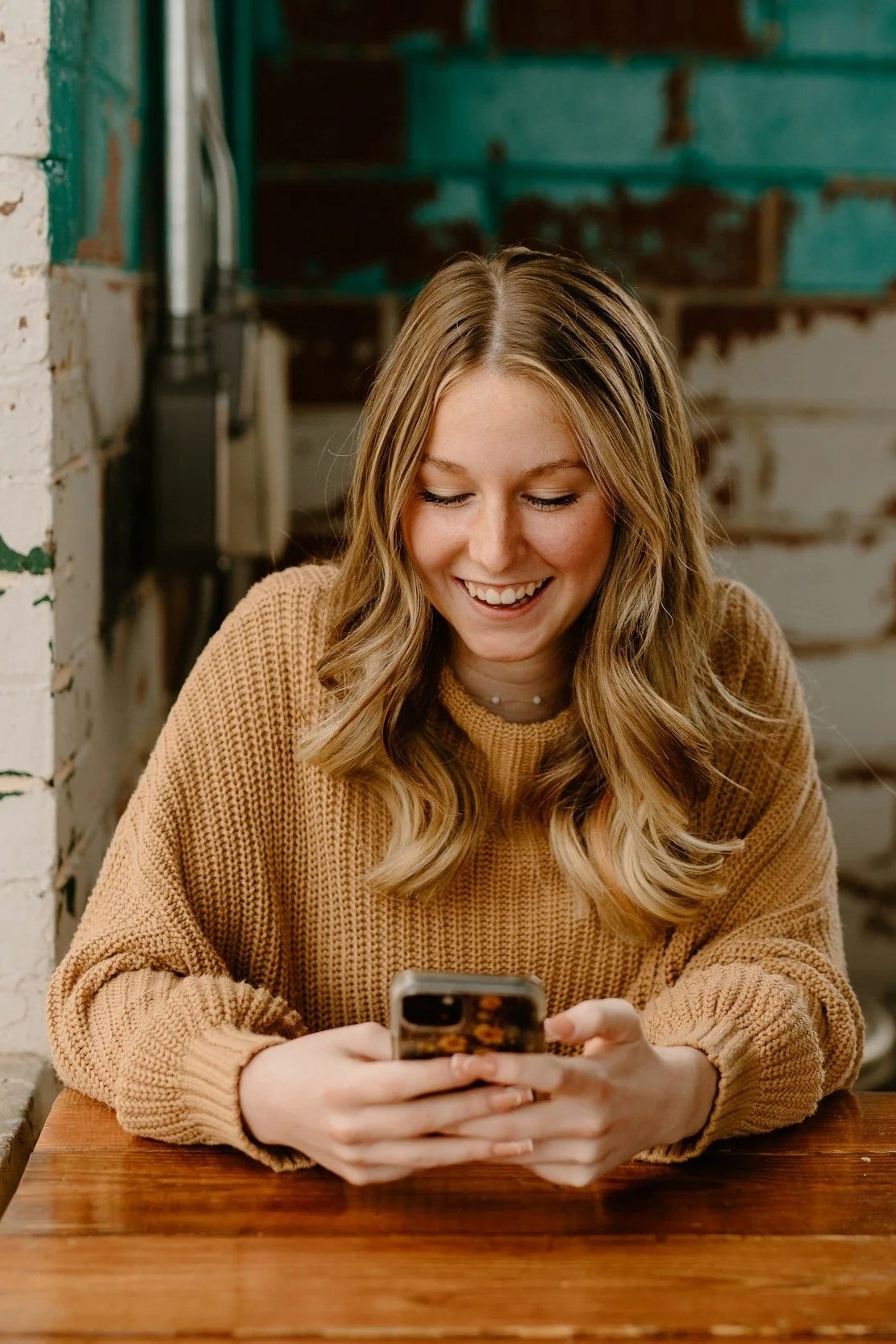 A young woman with wavy blonde hair wearing a tan sweater, sitting at a wooden table, smiling and looking at her phone.