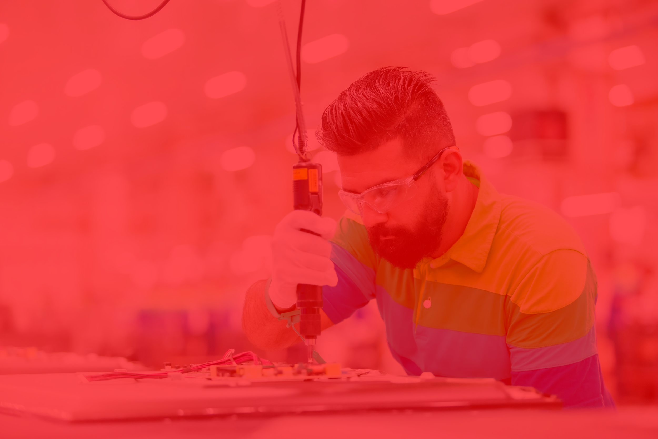 A worker wearing safety goggles and gloves, using a power drill on a flat panel in an industrial setting.