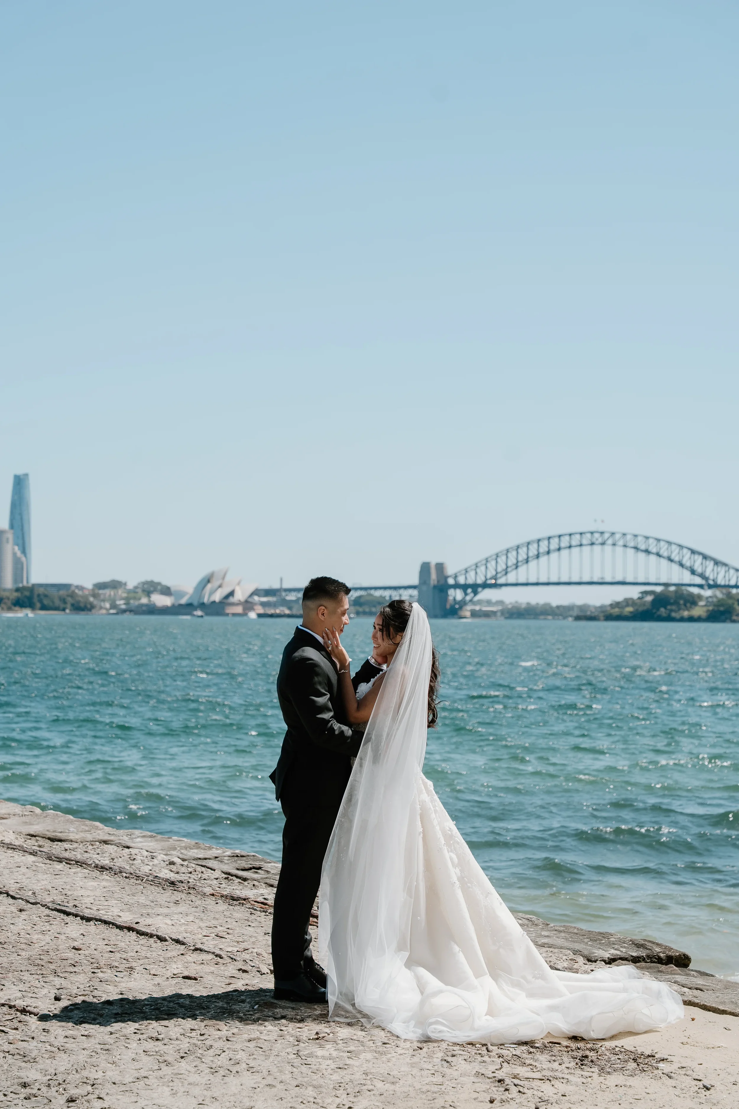 A bride and groom stand on a beach with the Sydney Harbour Bridge and Opera House in the background, sharing a romantic moment.