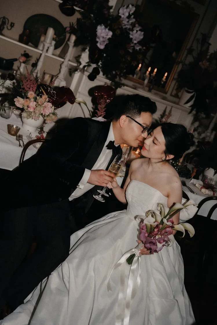 A bride holding a bouquet of calla lilies and purple flowers, and a groom in a tuxedo, sharing a moment at their wedding reception with champagne glasses.