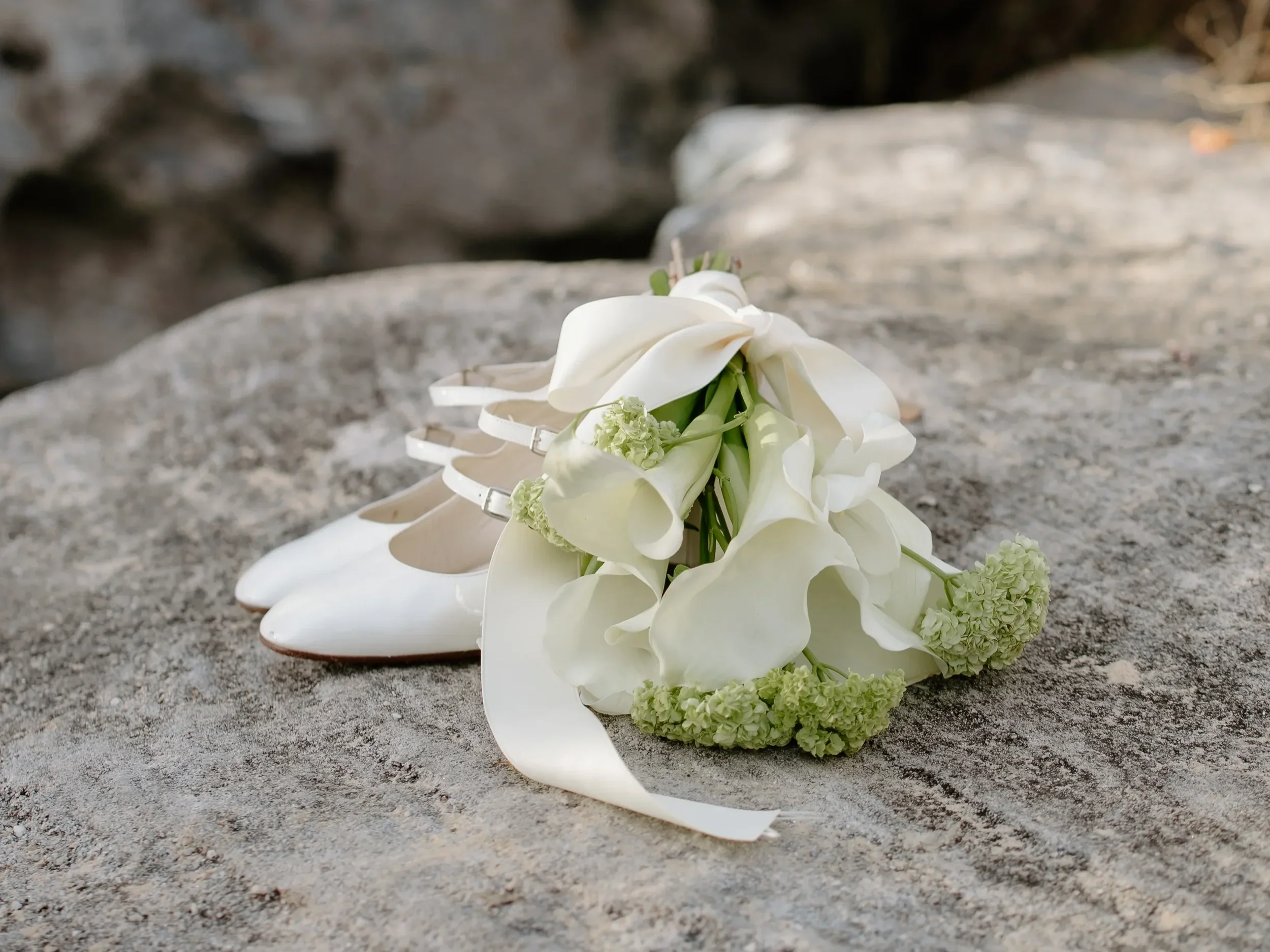 White wedding shoes and a bouquet of white flowers on a stone surface.