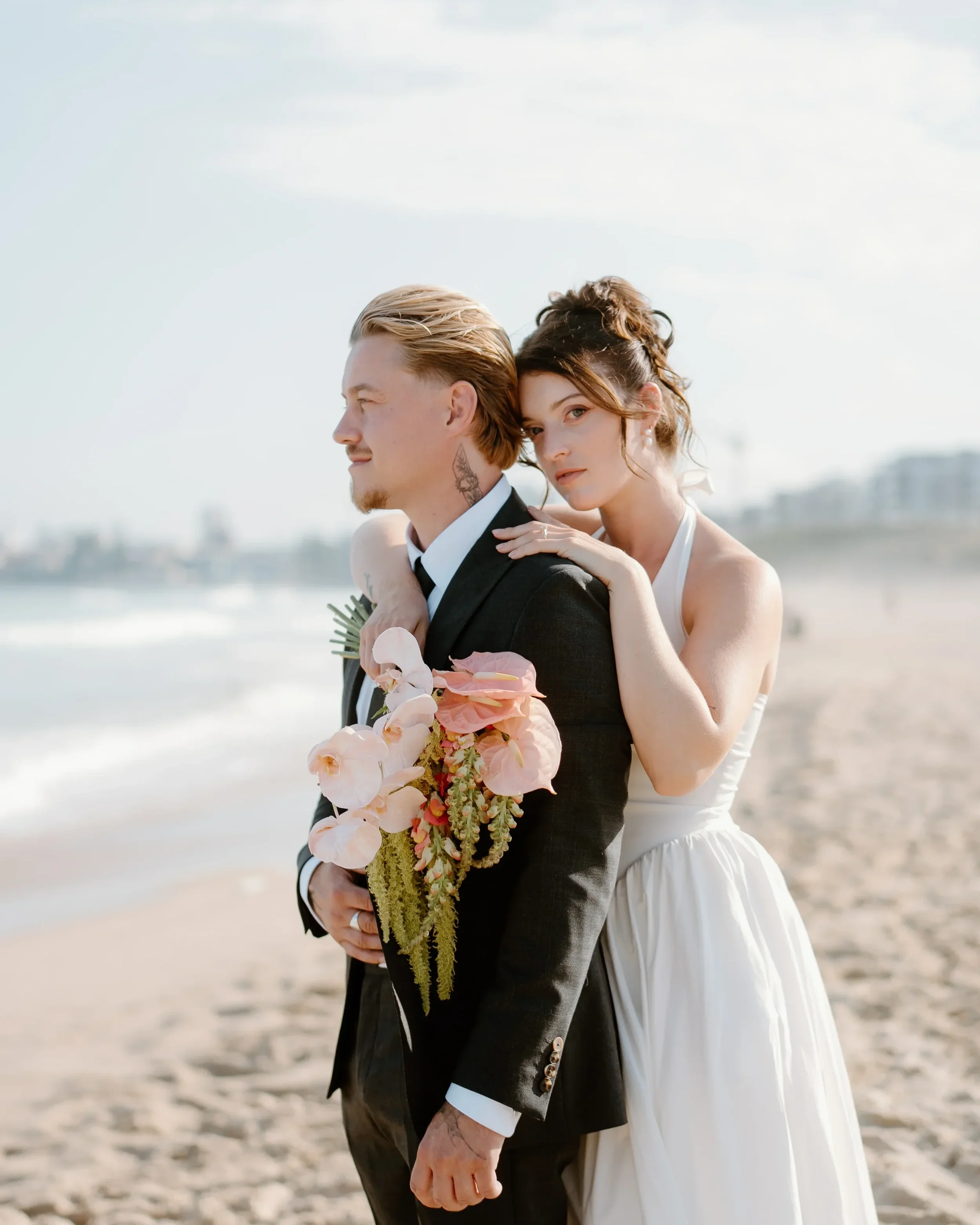 A couple in wedding attire on the beach, the man is holding a bouquet of pink flowers, and the woman is embracing him from behind with her hand on his shoulder.