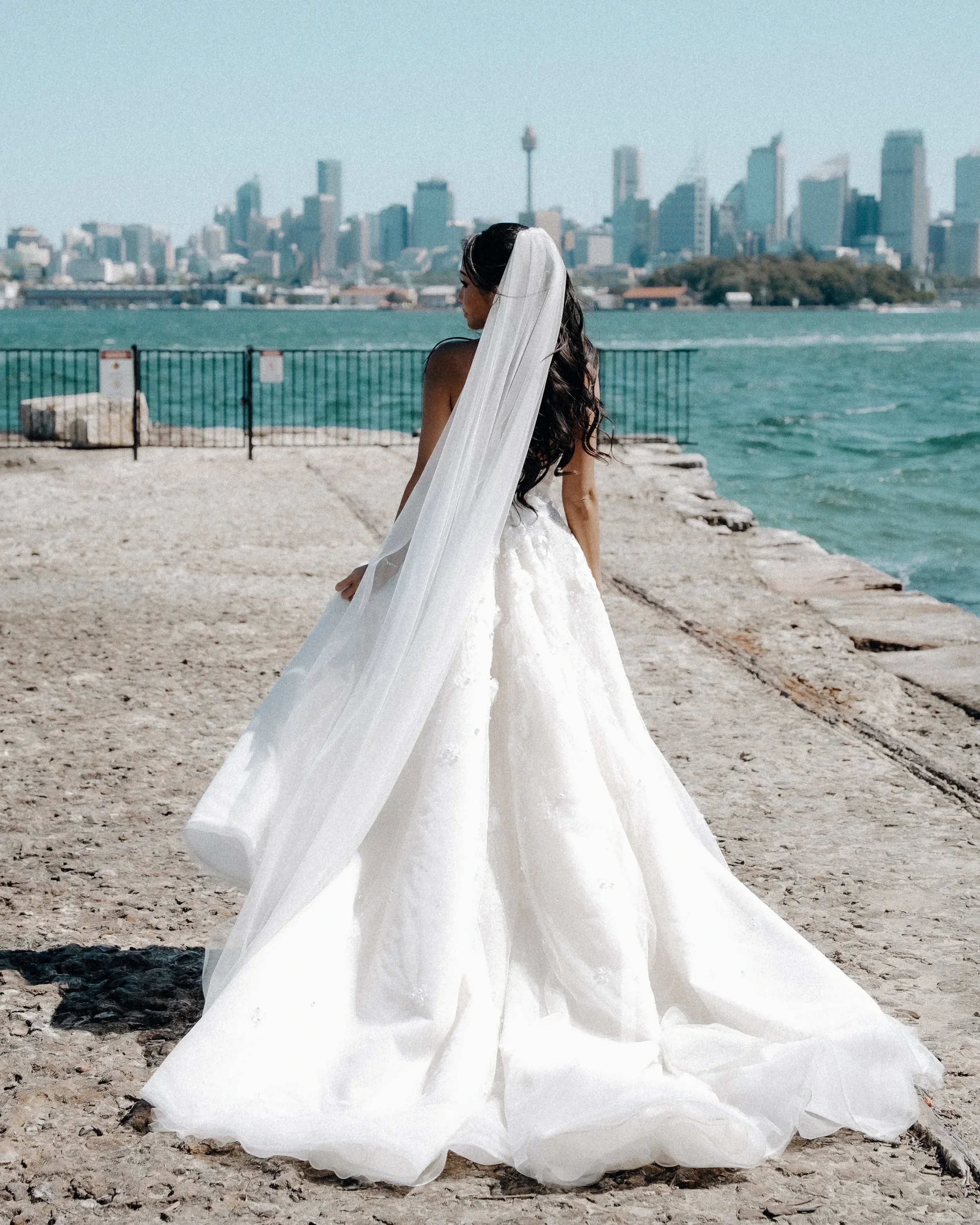 A woman in a white wedding dress with a long veil standing on a pier near the water with a city skyline in the background.
