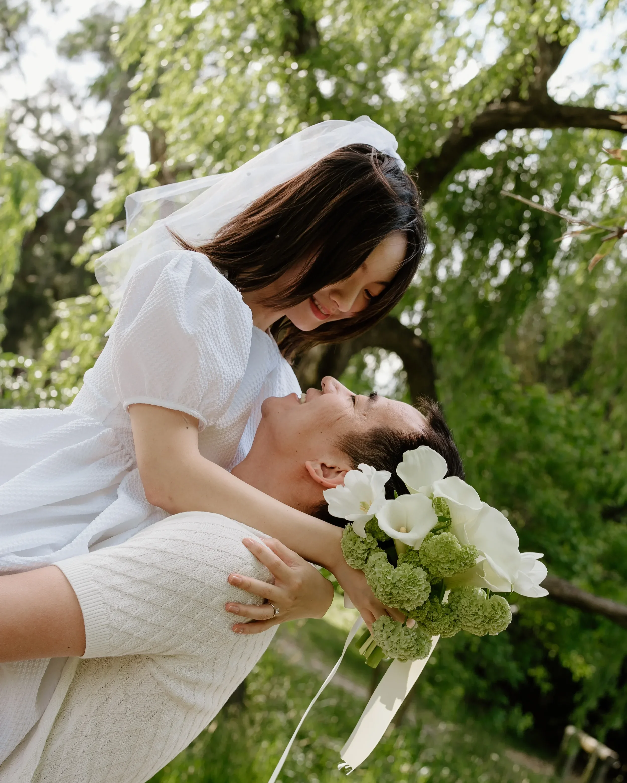 A couple sharing a joyful moment outdoors with trees in the background, one person holding a bouquet of white flowers with green accents.