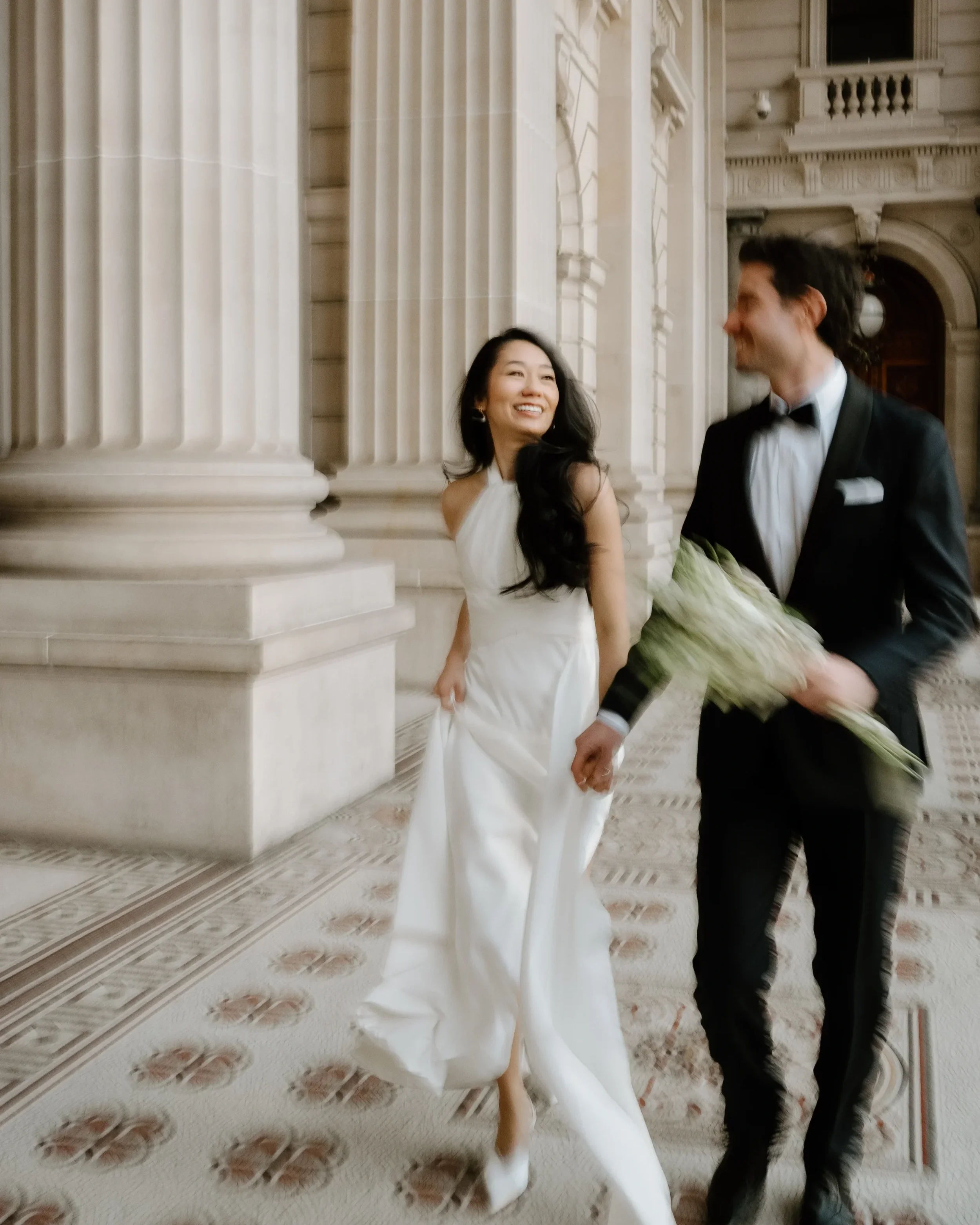A smiling woman in a white wedding dress standing next to a man in a black tuxedo holding a bouquet of flowers inside a grand, ornate building.