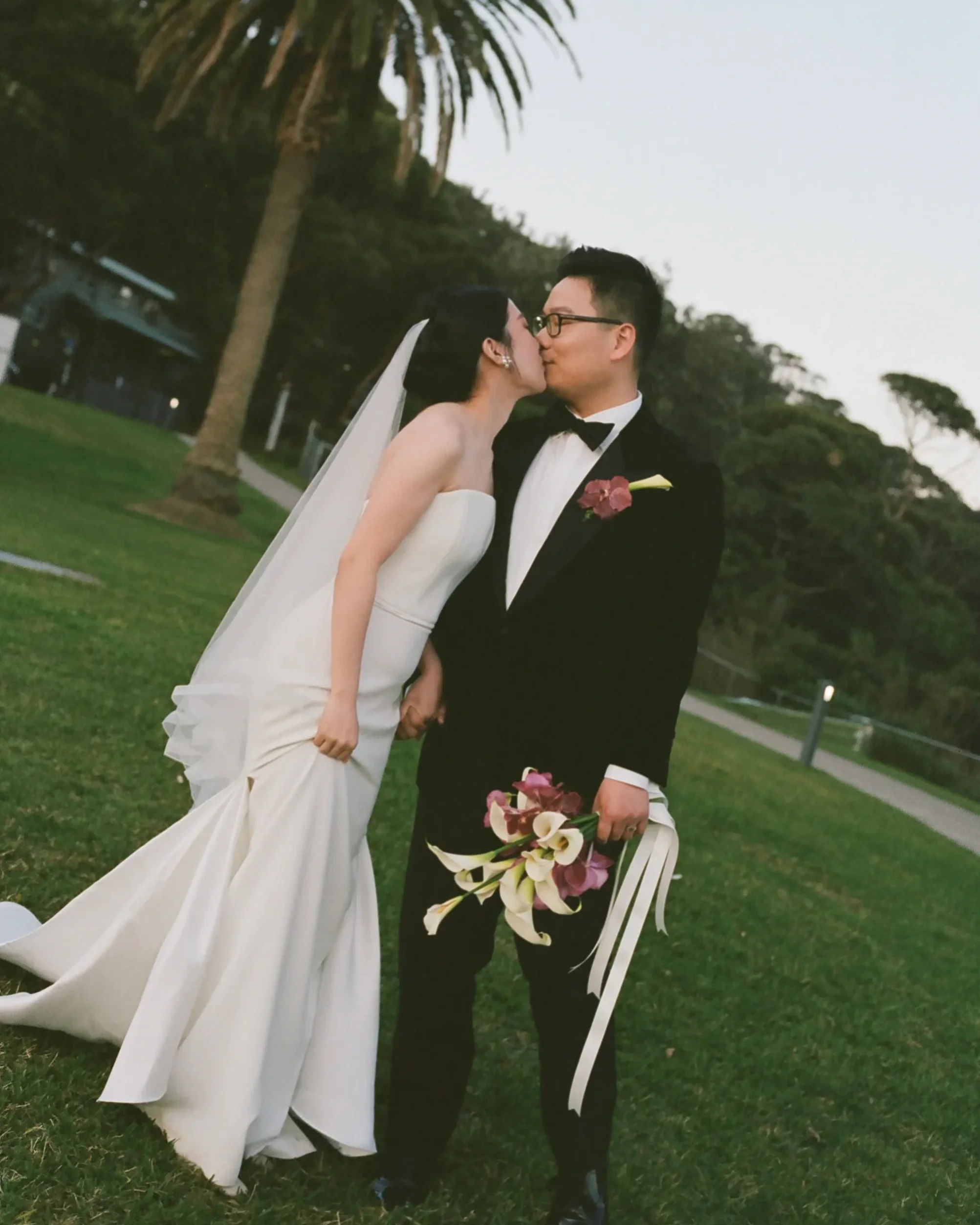 A newlywed couple, a woman in a strapless white wedding gown with a long veil and a man in a black tuxedo with glasses, are sharing a kiss outdoors on a grassy area with trees and a pathway in the background. The man holds a bouquet of white and pink