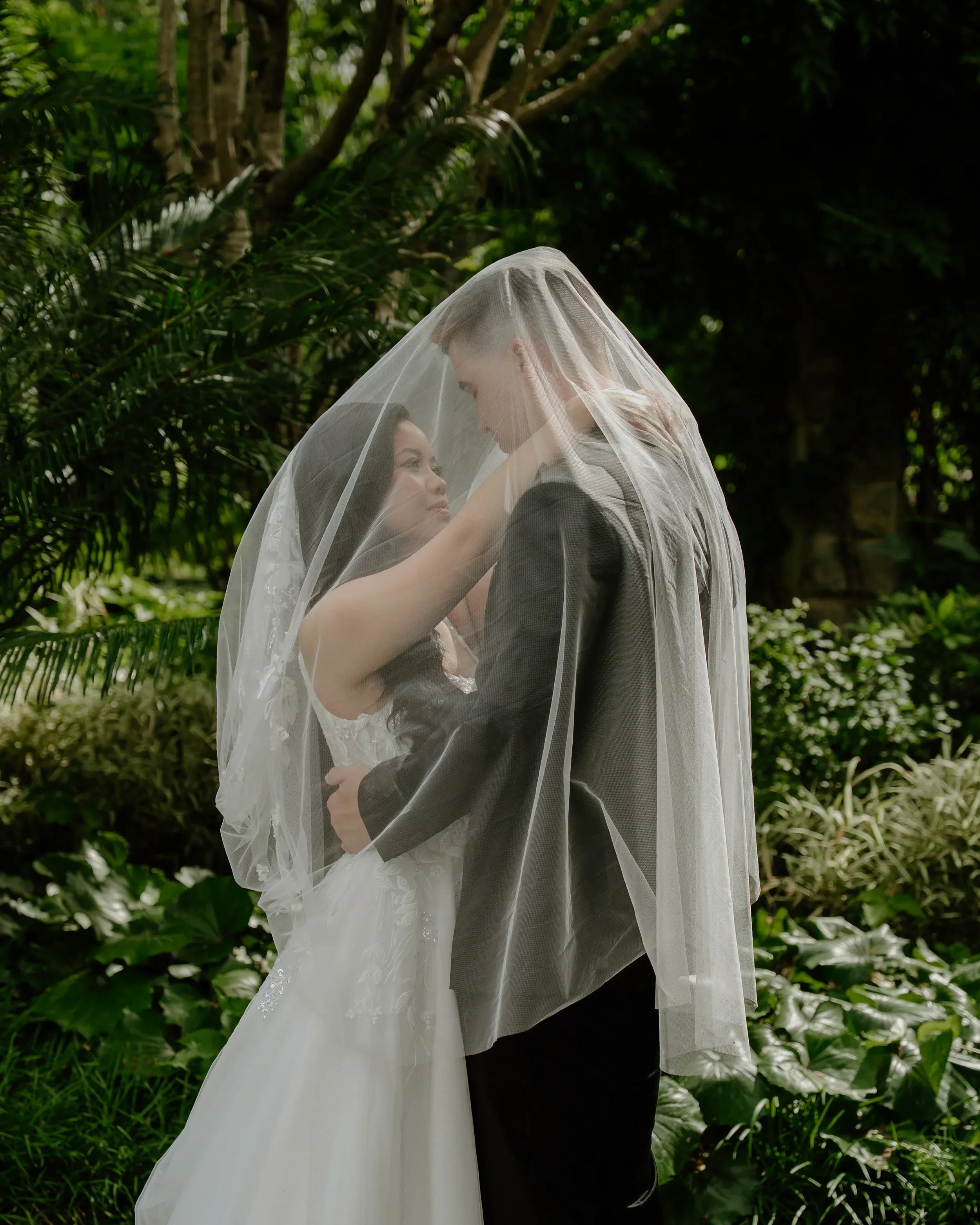 Bride and groom under bridal veil in a lush outdoor garden setting.