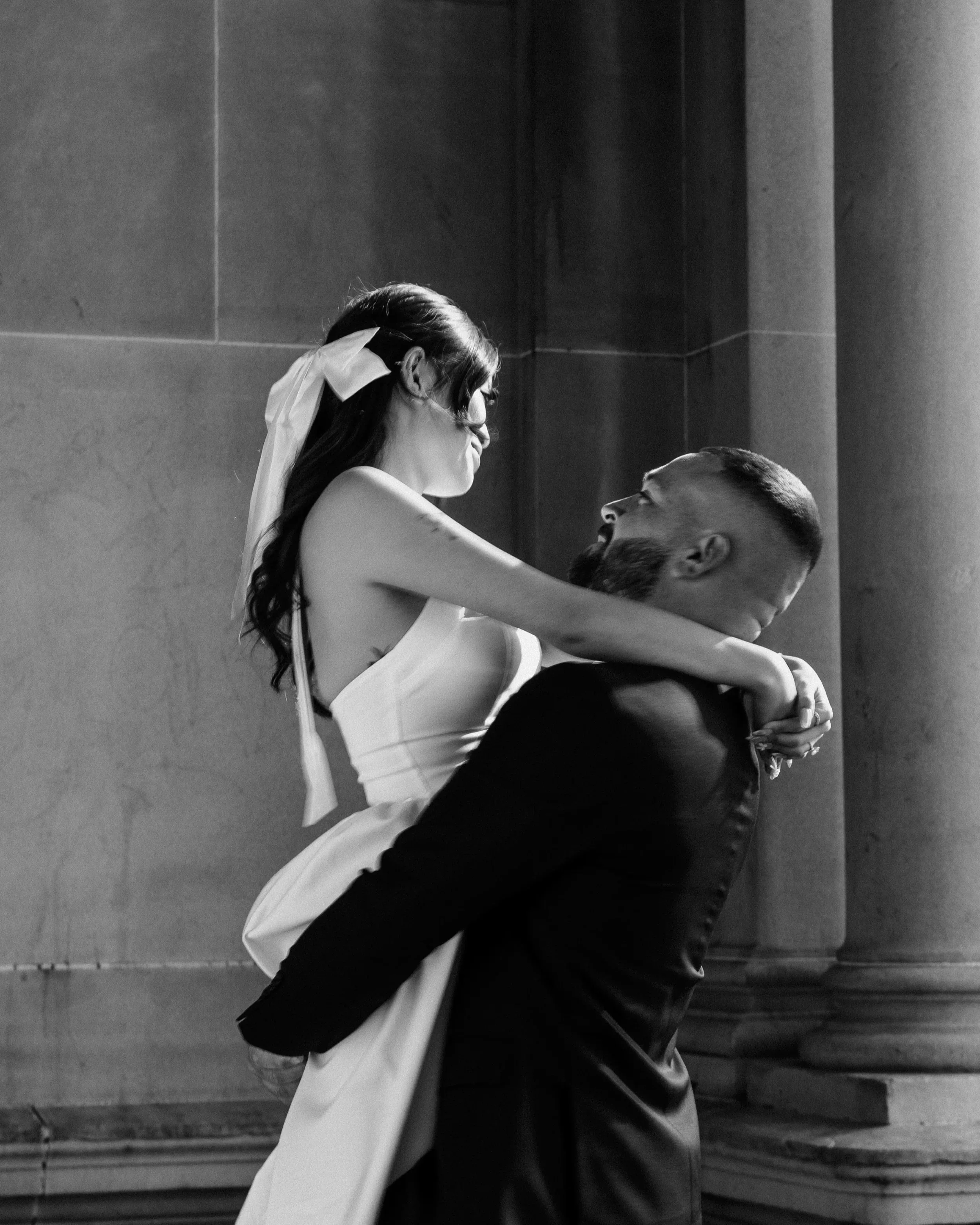 A man in a suit lifts a woman in a wedding dress, both smiling and looking at each other, in a black and white photo.
