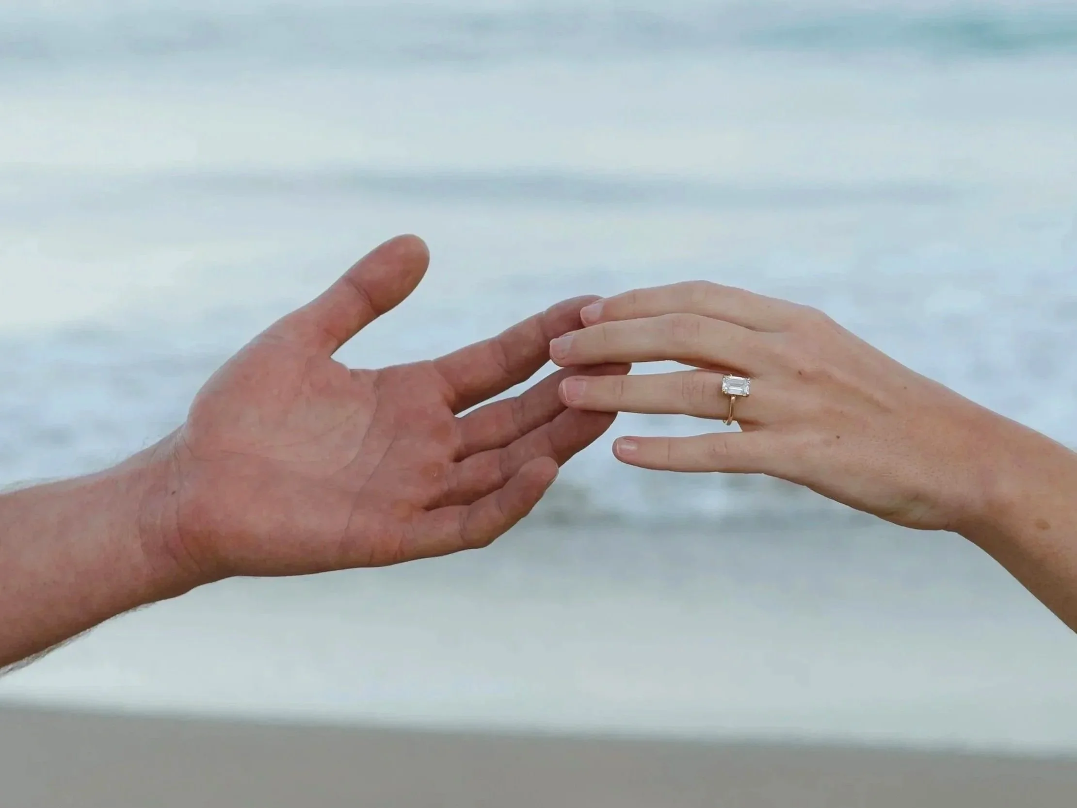 Hands reaching towards each other with a focus on one person's engagement ring, against a beach background.