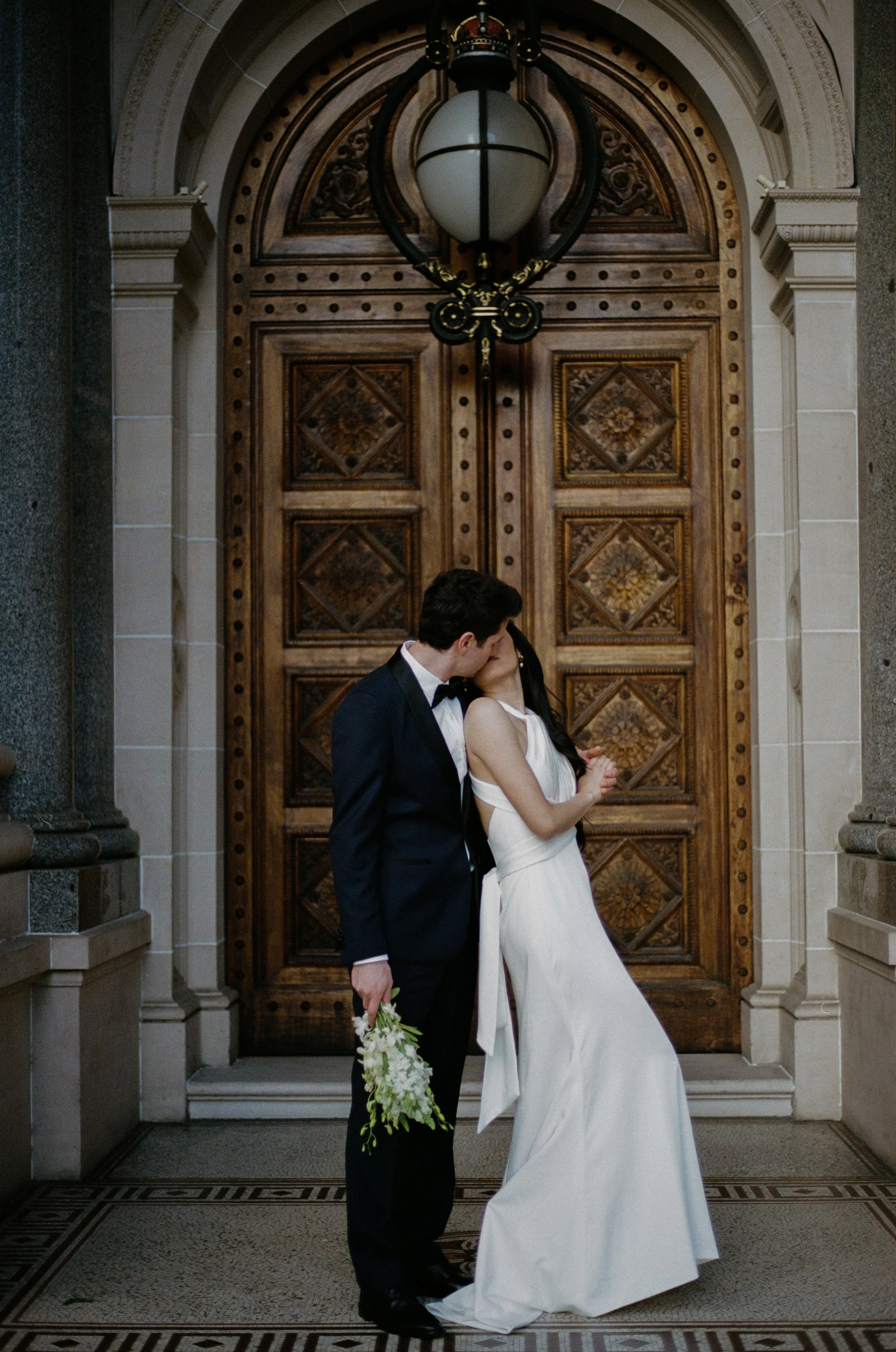 A bride and groom share a kiss in front of a large, ornate wooden door with intricate carvings. The groom is in a dark suit and white shirt, holding a bouquet of white flowers. The bride is in a white wedding gown, leaning into the groom.