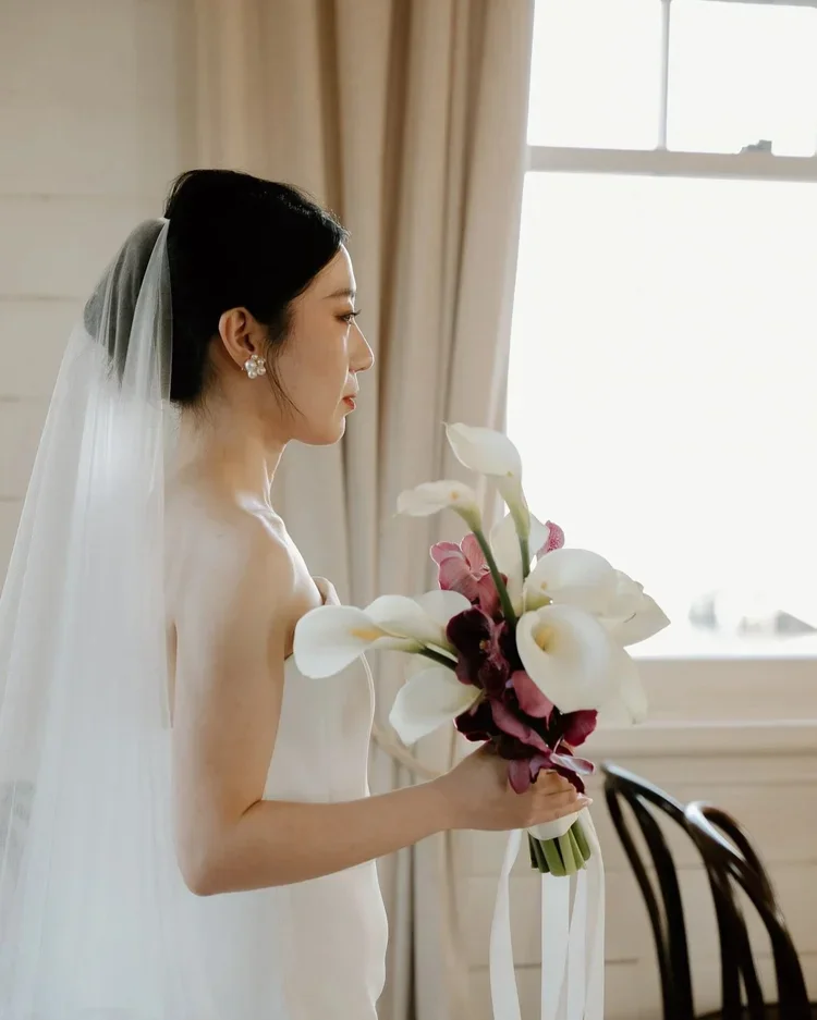 Bride in a wedding dress holding a bouquet of white calla lilies and pink flowers, standing indoors near a window with beige curtains.