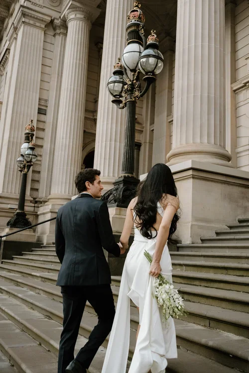 A bride and groom standing on the steps of a grand, classical building with large columns, holding hands. The bride has long black hair, is wearing a white gown, and holding a bouquet of white flowers. The groom is dressed in a dark suit. Vintage-style street lamps are visible in the background.