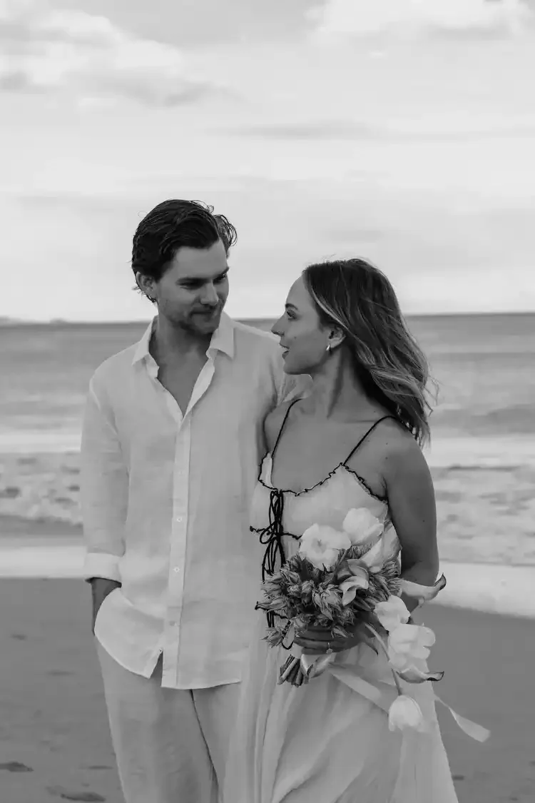 A black-and-white photo of a man and woman walking on the beach together. The woman is holding a bouquet of flowers and looking at the man. The man is looking at the woman. The scene appears romantic and serene.