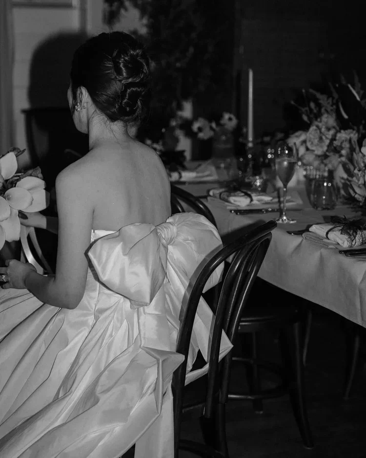 A woman in an elegant strapless gown with a large bow on the back, sitting at a dining table set for a formal event.