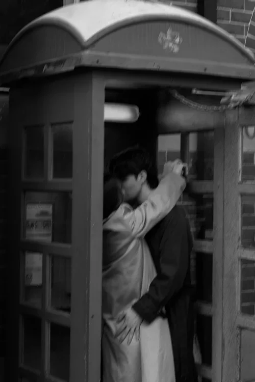 Two people kissing inside a small, vintage-style phone booth near a brick wall.