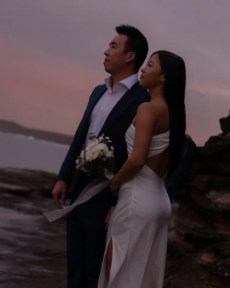 A couple dressed in formal attire standing close on a beach during sunset, the man in a suit holding a bouquet of flowers, both looking into the distance.