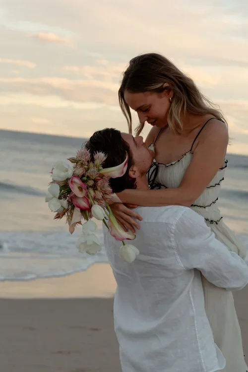 A couple on the beach, with the woman holding the man and smiling, with a floral crown on the man's head and the ocean in the background at sunset.