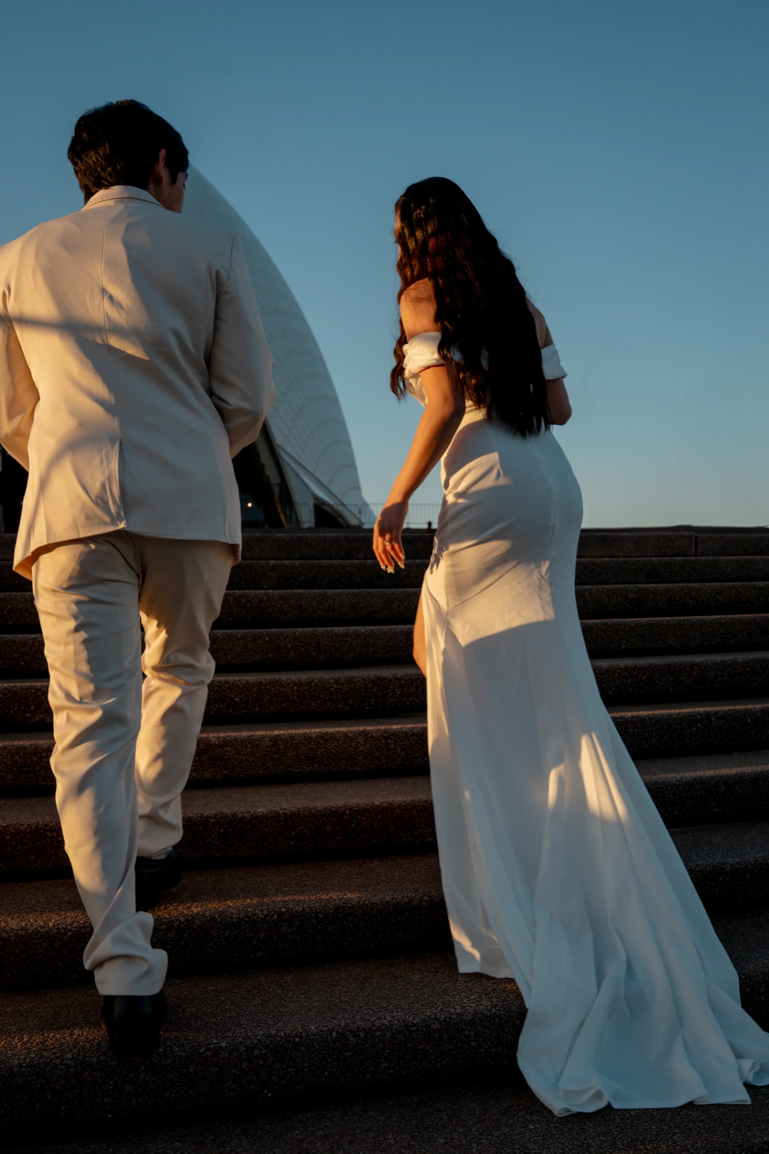 A woman in a white wedding dress and a man in a light-colored suit ascending outdoor stairs at sunset.
