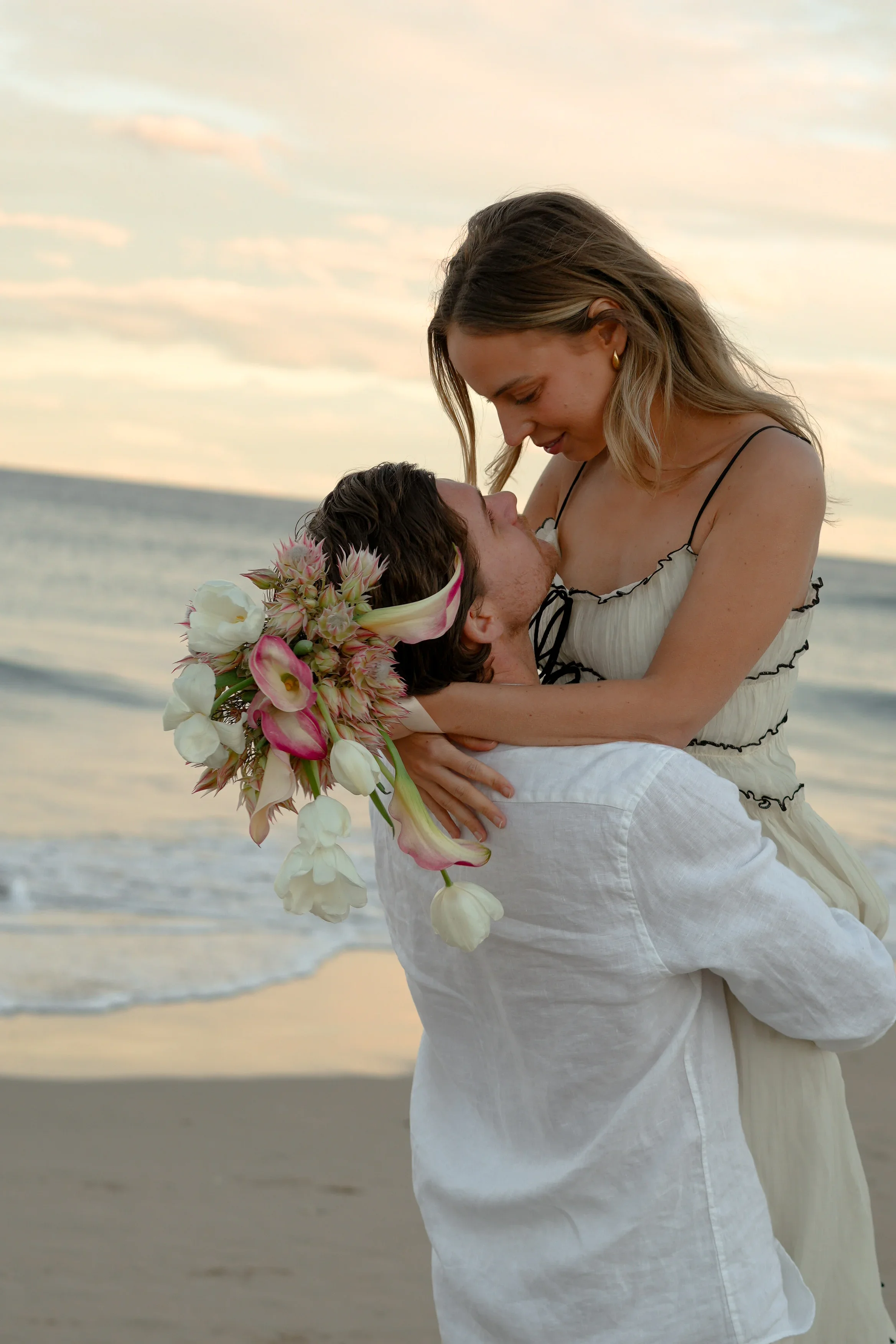 A couple embracing on the beach at sunset; the woman is wearing a dress with spaghetti straps and gold earrings, the man is holding a large bouquet of flowers with white and pink blooms, and they are gazing at each other.