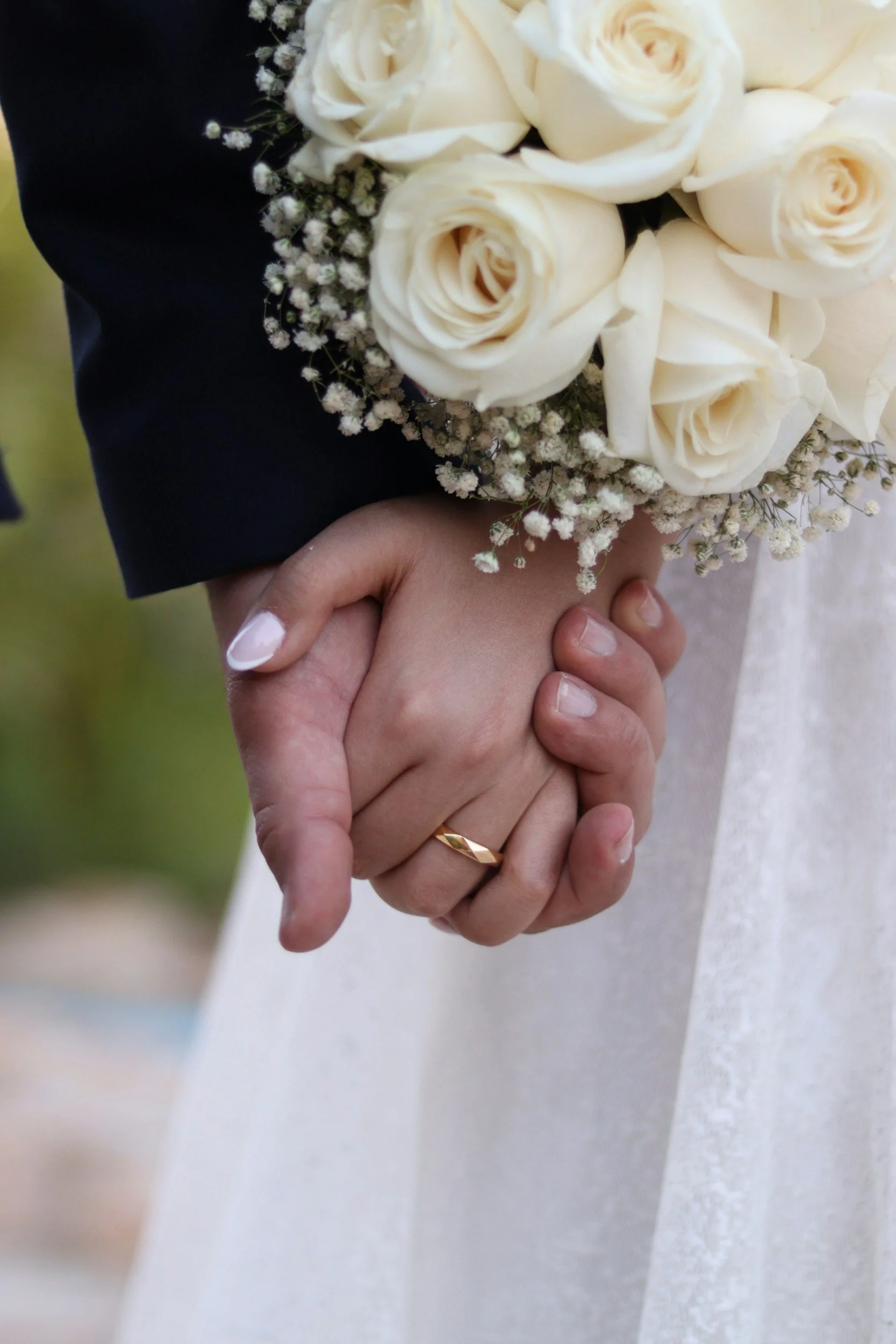 Close-up of two people holding hands, with one wearing a gold wedding ring and a white wedding dress, and the other person wearing a black suit jacket, holding a bouquet of white roses and baby's breath.