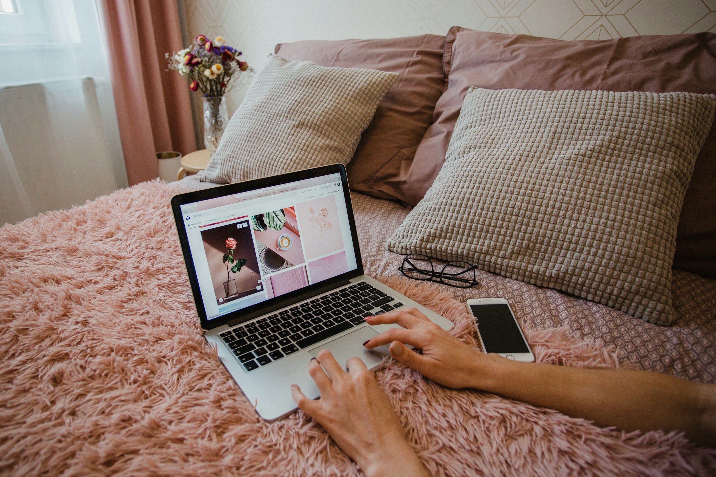 Person using a laptop on a pink furry blanket in a bedroom, with pillows, a bedside table, a vase with flowers, and a smartphone nearby.