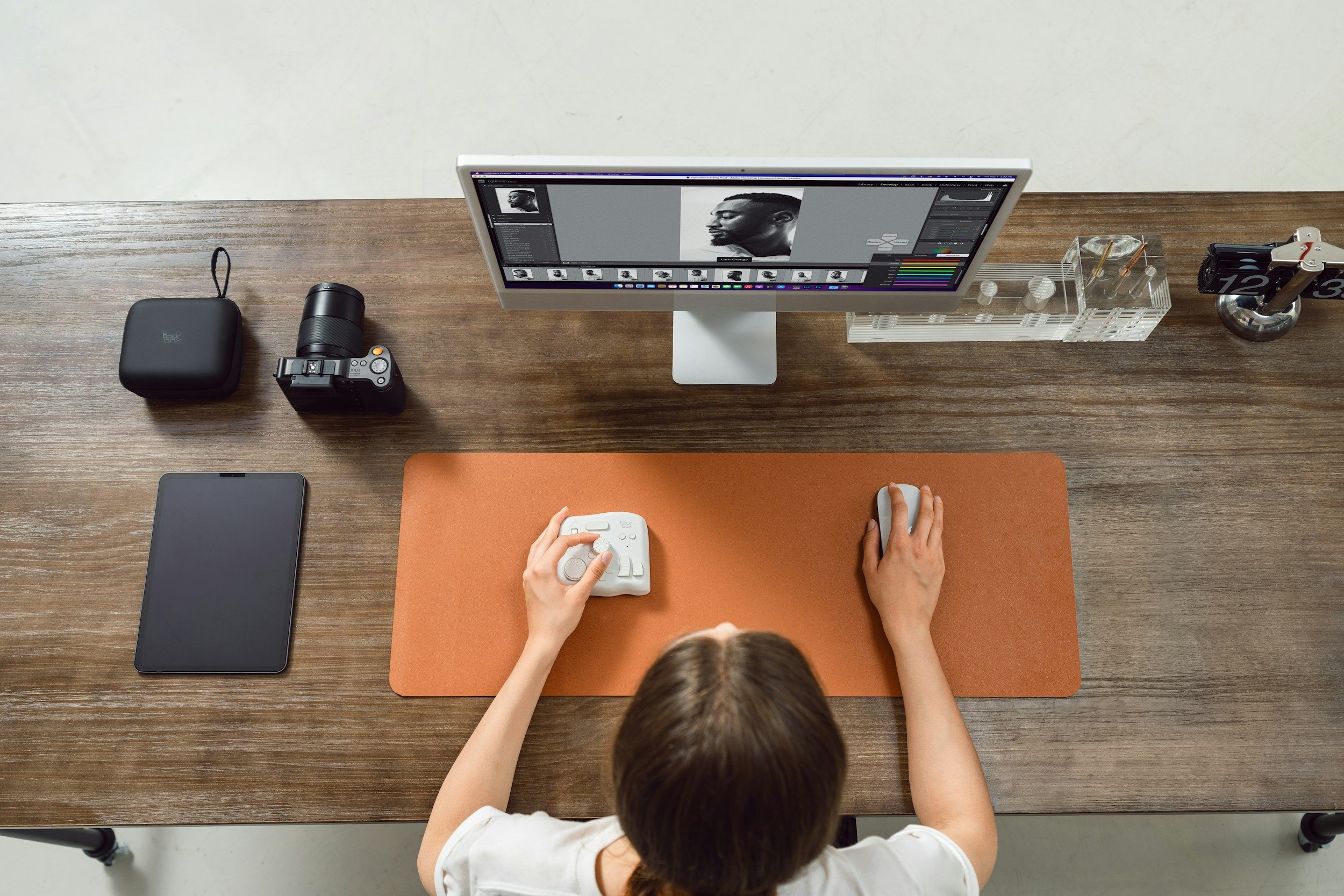 A person with brown hair in a white shirt using a mouse and keyboard at a wooden desk with a brown mouse pad. There is a tablet on the left, a camera with a strap, a computer monitor displaying black-and-white profile photo editing software, a transparent container with small items, and a cylindrical caliper and object holder.