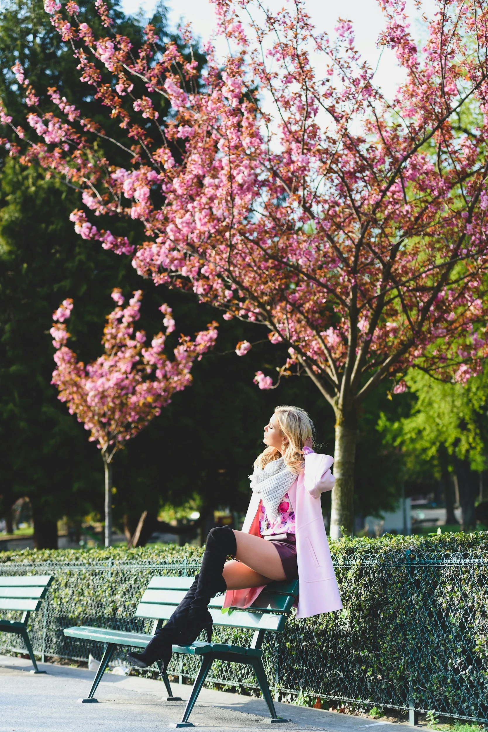A young woman with blonde hair sitting on a park bench beneath pink flowering trees. She is wearing a pink blazer, a light-colored scarf, a floral blouse, and black thigh-high boots. She is looking away from the camera with a relaxed expression in a sunny outdoor setting.