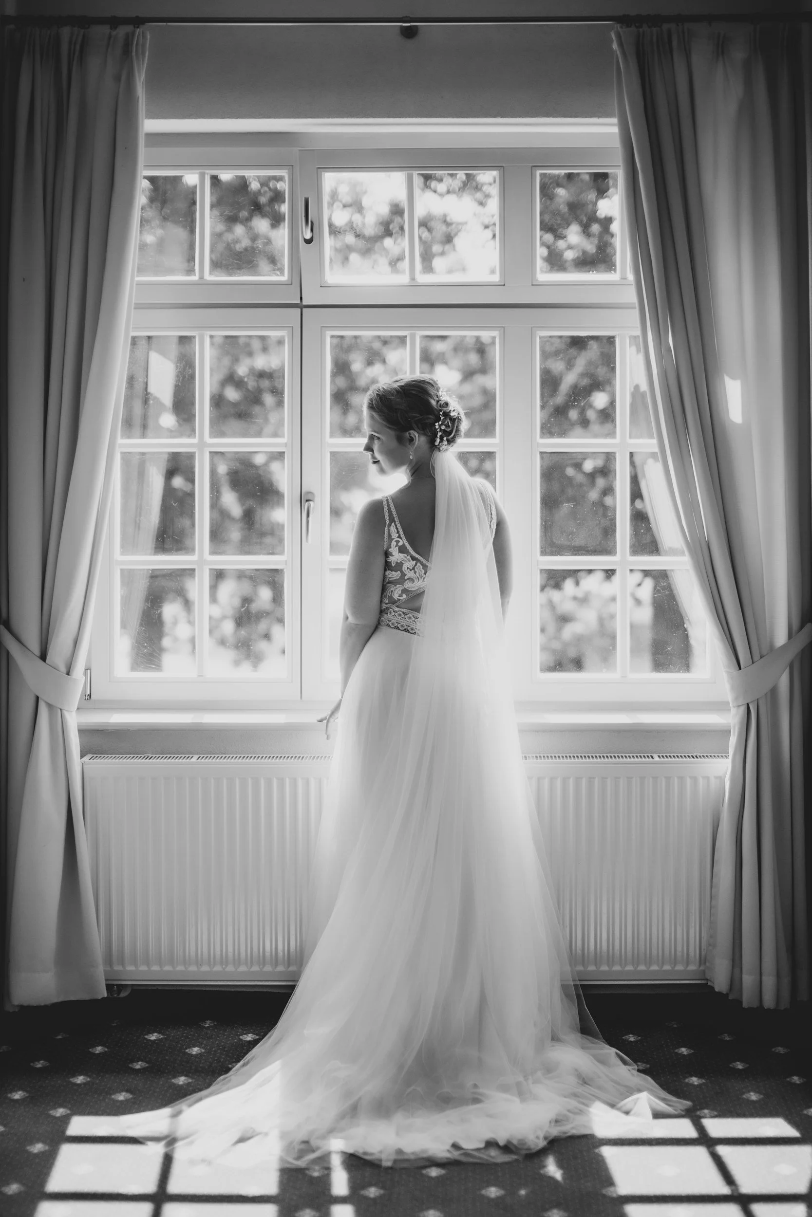 A bride in a wedding dress standing by a large window with curtains, looking outside, in black and white photograph.