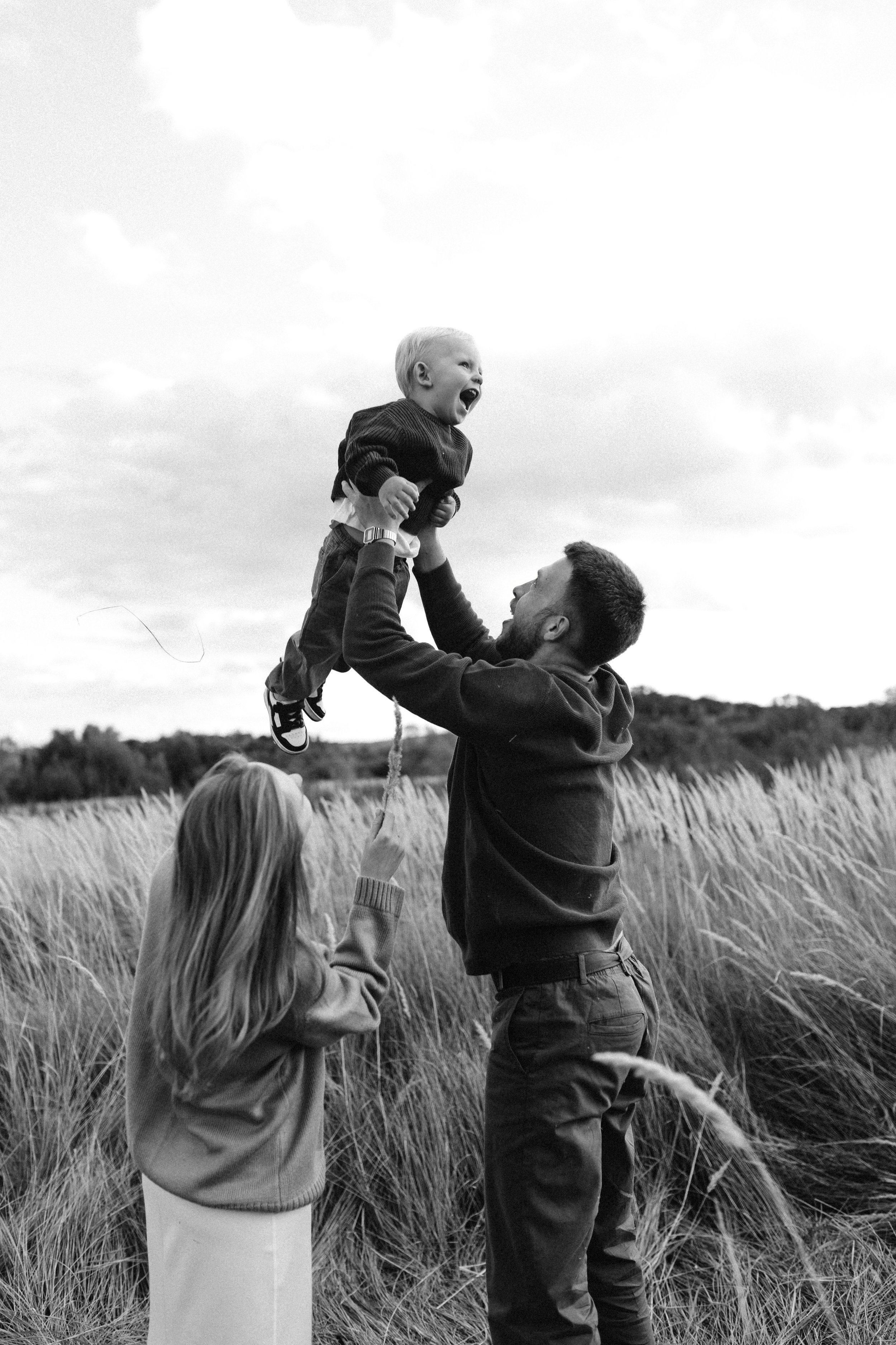 A man lifting a smiling young boy in the air outdoors in a field of tall grass, with a woman standing nearby and pointing, all in black and white.