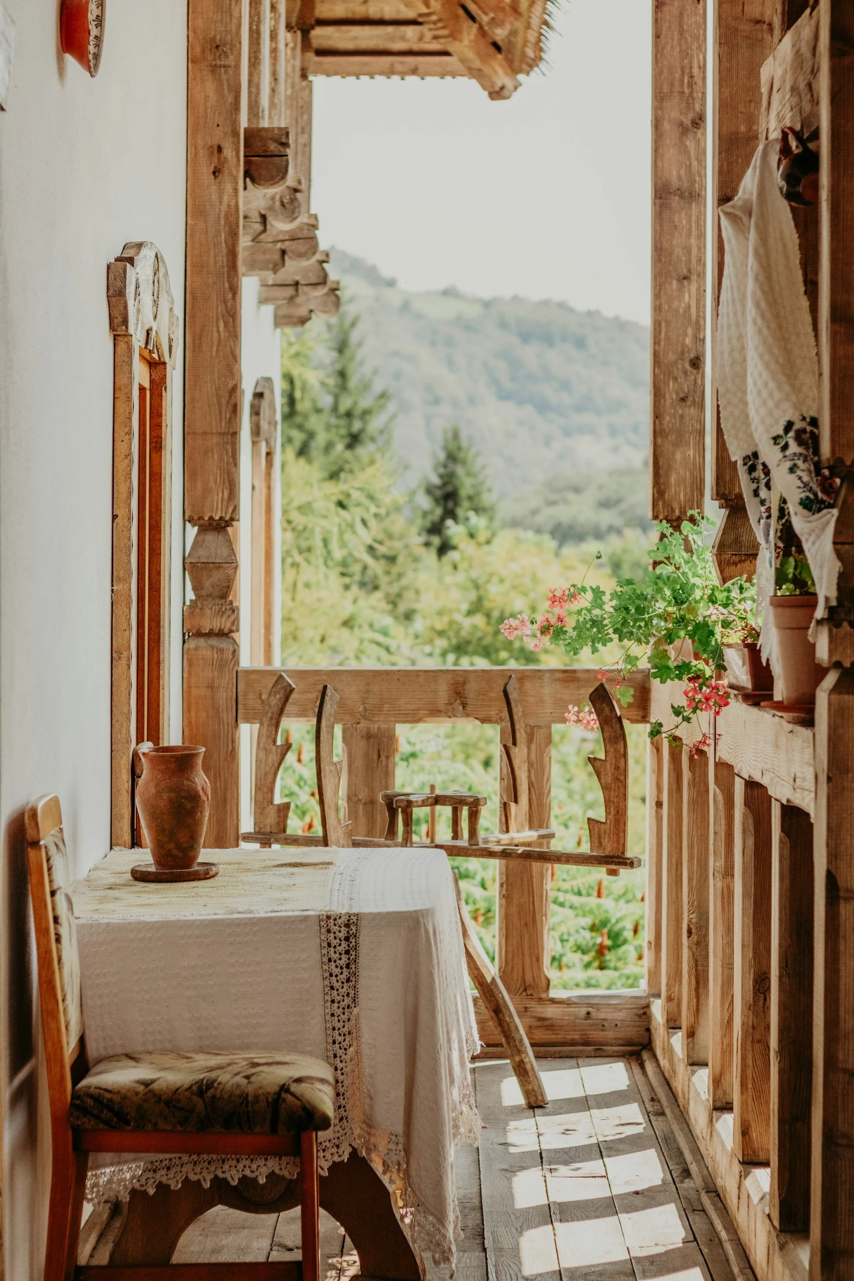 A rustic balcony with a small wooden table covered with a white lace tablecloth, a potted clay vase, and a chair with a floral cushion. A wooden railing surrounds the balcony with potted pink flowers, and a scenic view of trees and hills in the background.