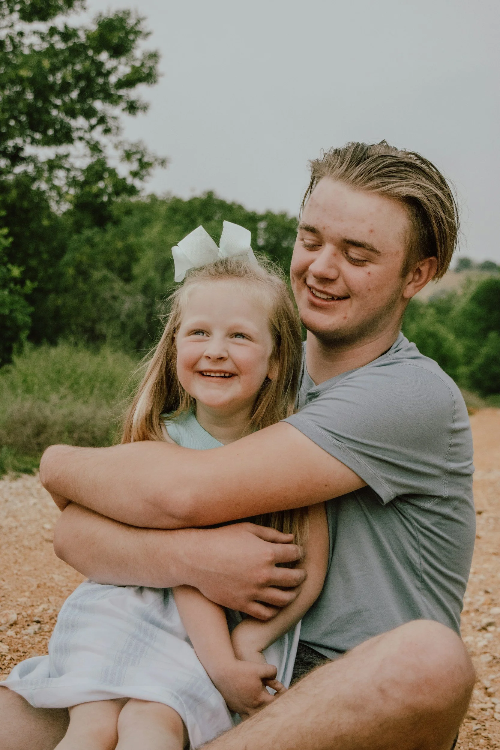 A young man and a little girl are sitting outdoors on a dirt path, hugging and smiling happily. The girl has a white bow in her hair and is wearing a light blue top with a white dress. The man has light brown hair, is wearing a gray t-shirt, and has his eyes closed as he smiles.