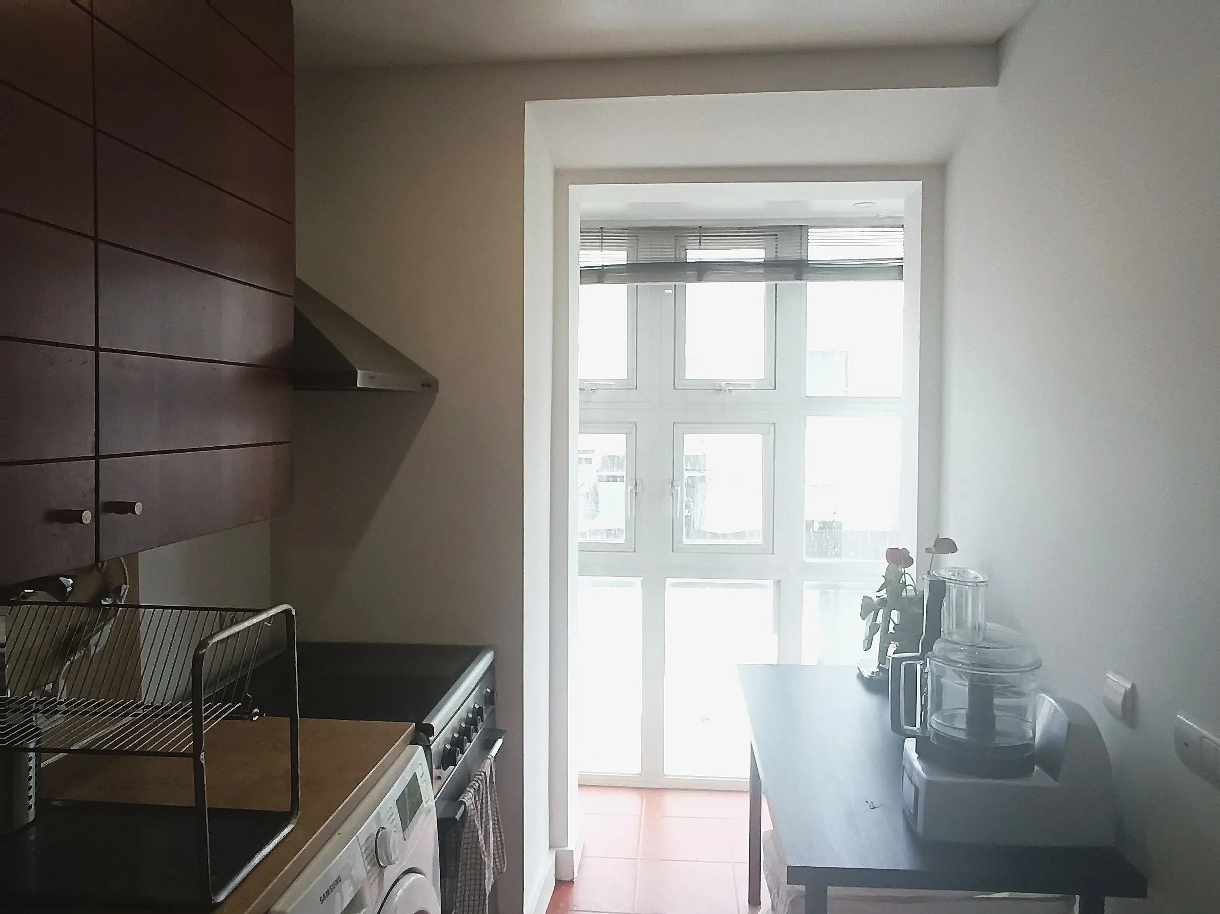 Small kitchen with wooden cabinets, a stove, a washing machine, a counter with a dish rack, and a table with a blender and a vase with a flower. A large window with multiple panes is in the background.
