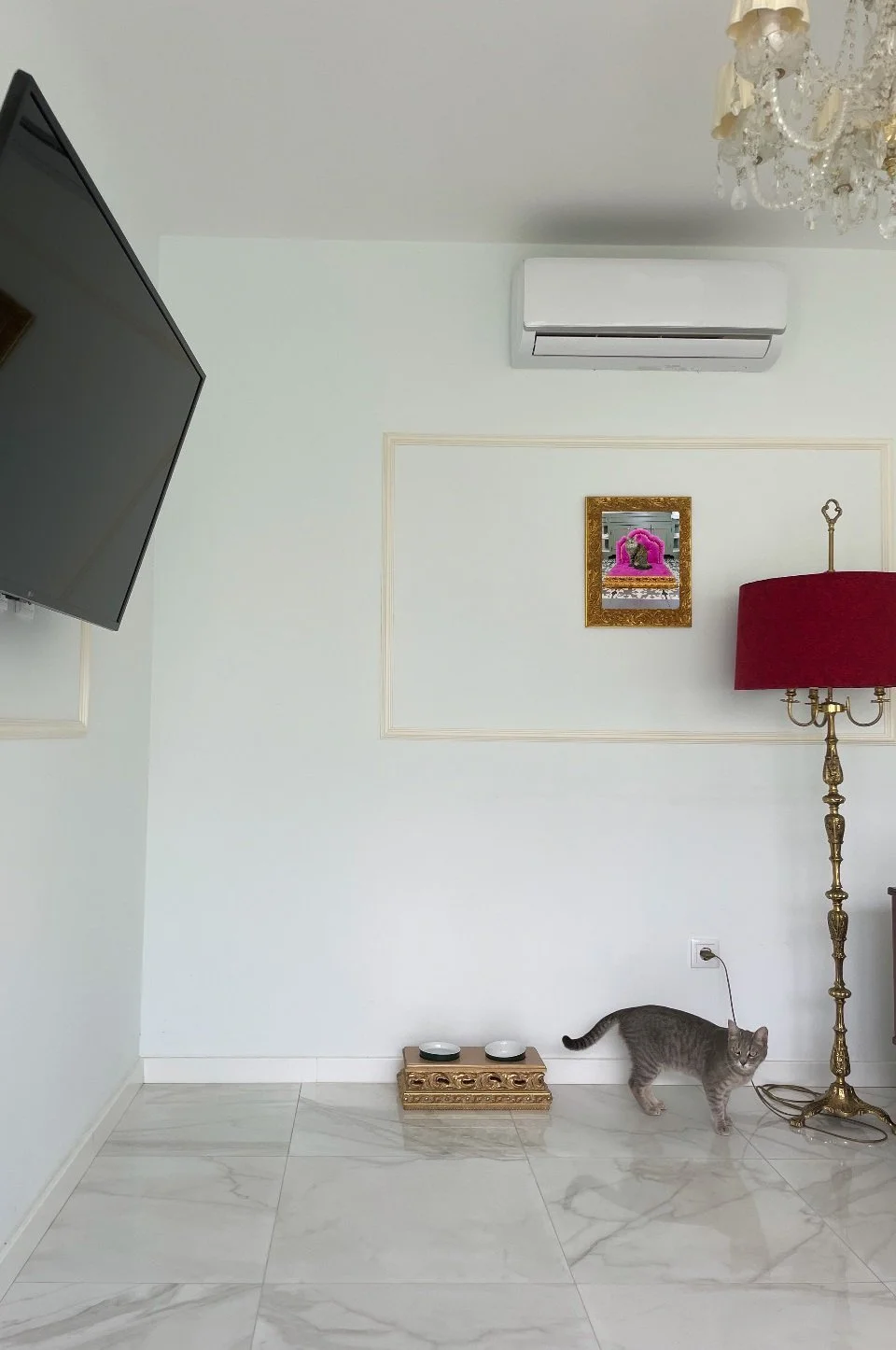 Interior living room with white walls and marble floor, featuring a mounted flat-screen TV, a wall air conditioning unit, a decorative picture frame with pink decorations, a red lampshade on a tall ornate lamp, a grey cat near a gold food bowl stand, and a chandelier hanging from the ceiling.