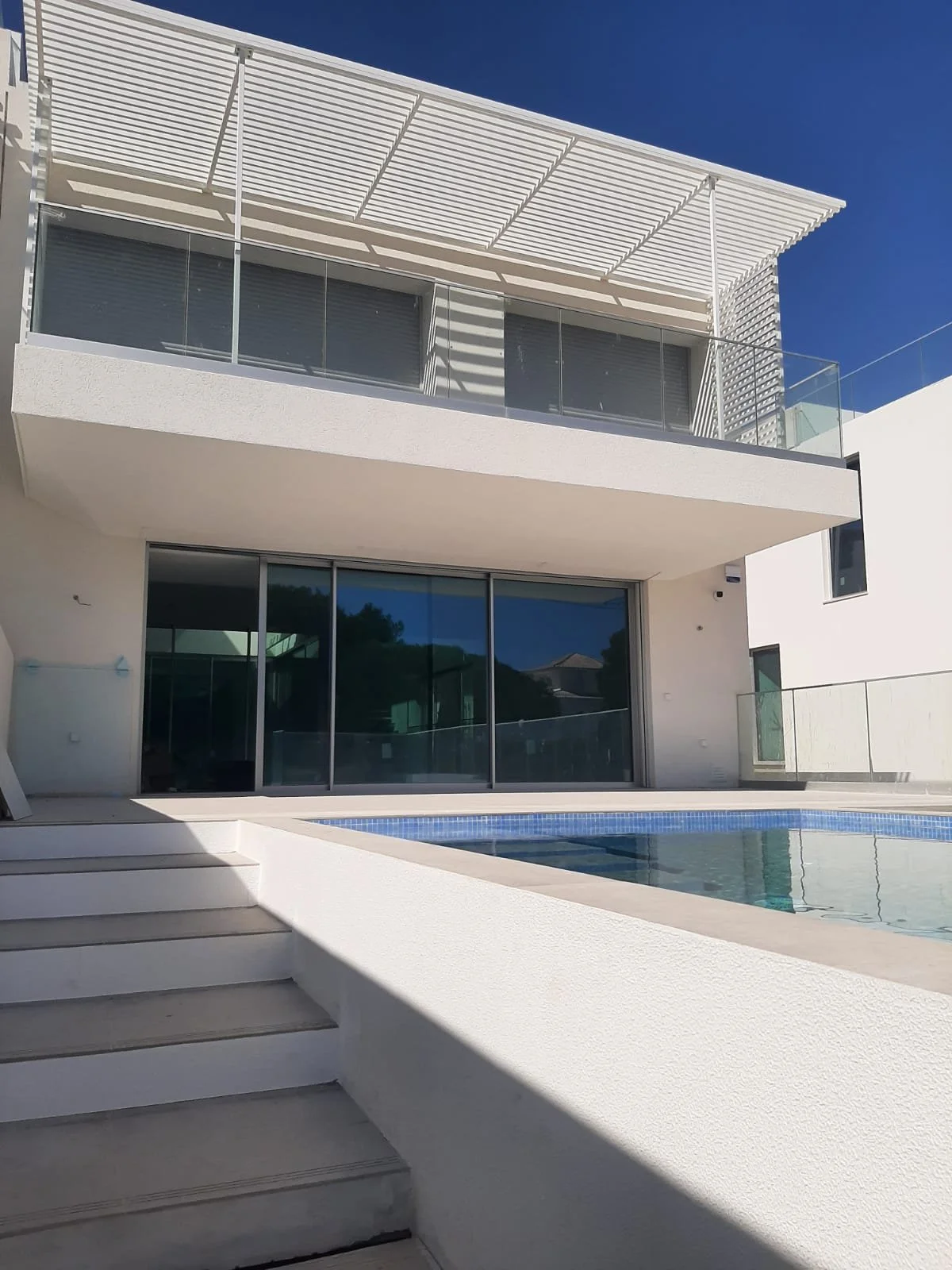 Modern white house with large glass sliding doors, a swimming pool, and a balcony with glass railings under a clear blue sky.