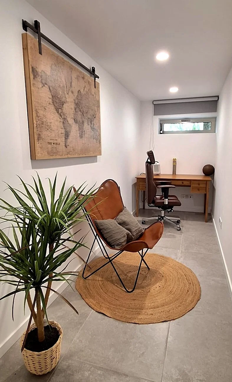 A small, modern home office with a desk, office chair, decorative globe, and window; a butterfly chair with pillows, a potted plant on a woven basket, a world map on the wall, and a round woven rug on gray tile floor.