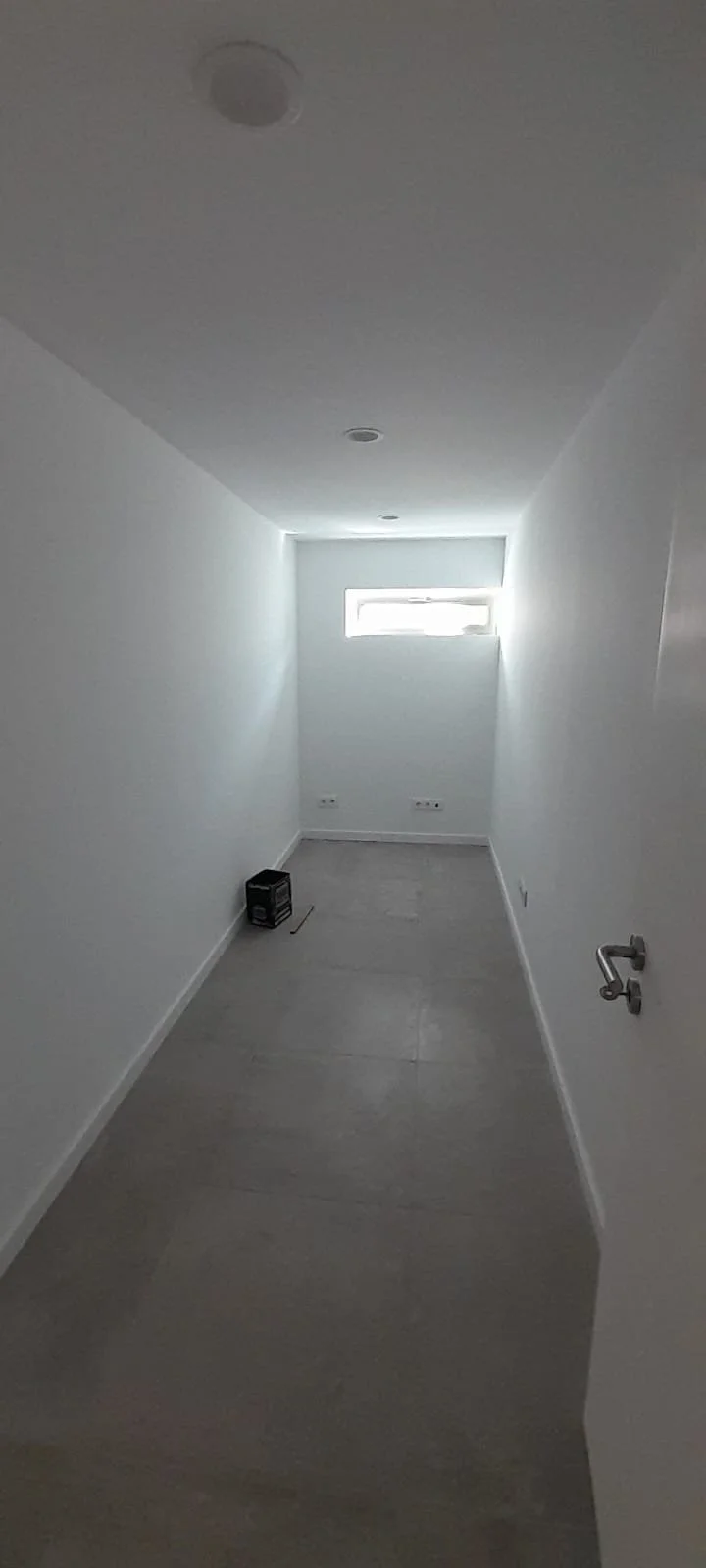 Empty narrow room with white walls, gray flooring, a small window near the ceiling, and a black object on the floor.