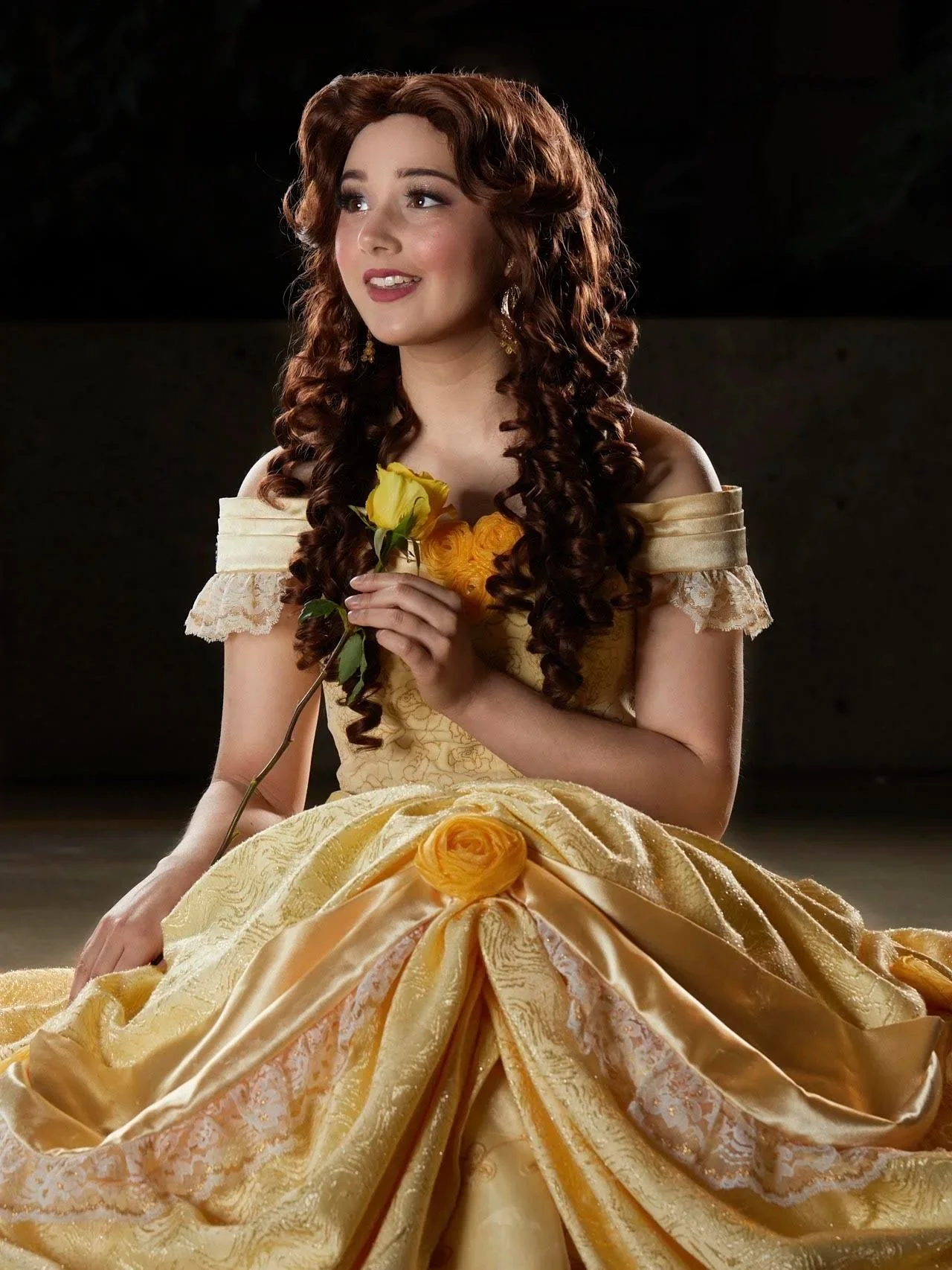 A woman dressed as Belle from Disney's Beauty and the Beast in an elaborate yellow ball gown with lace and floral details, holding a yellow rose, and sitting on the floor with a dark background.