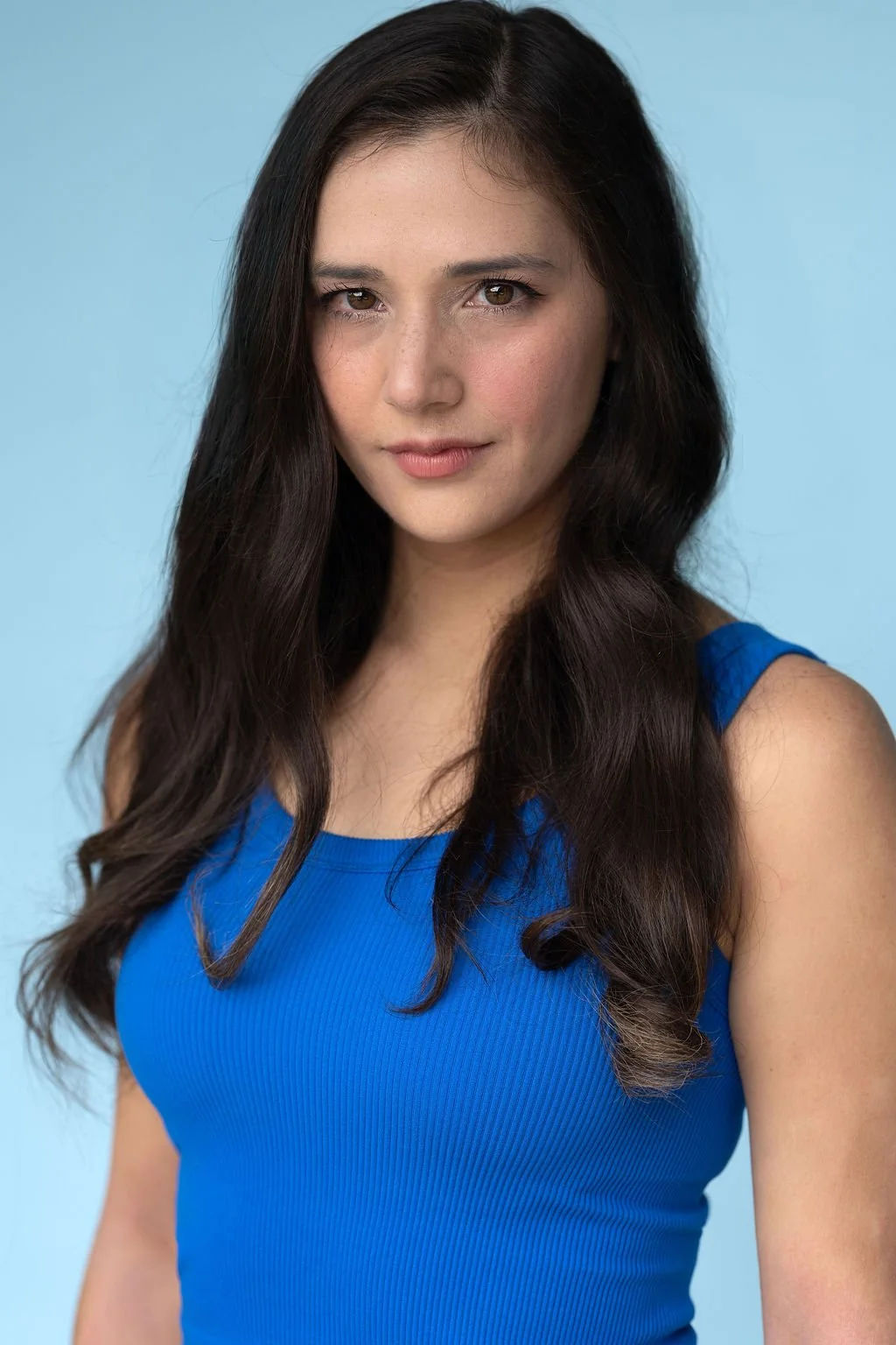 Young woman with long dark hair wearing a blue sleeveless top, looking at the camera with a slight smile, against a light blue background.