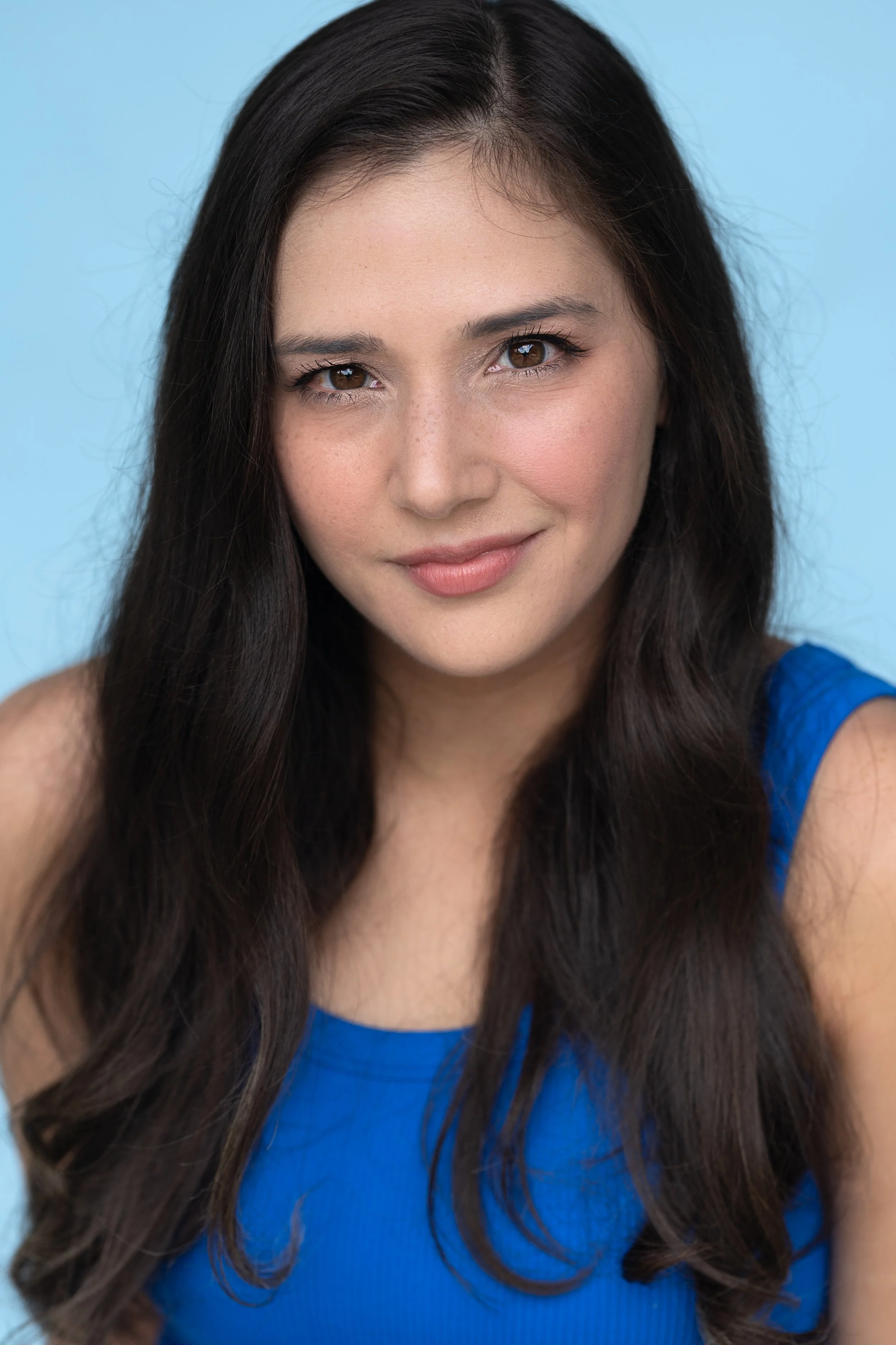 A portrait of a woman with long, dark brown hair wearing a blue sleeveless top, smiling softly against a light blue background.
