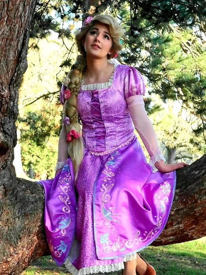 A woman dressed as Rapunzel from Disney's Tangled at a festival with colorful blurred lights in the background, smiling with her hands on her hips.