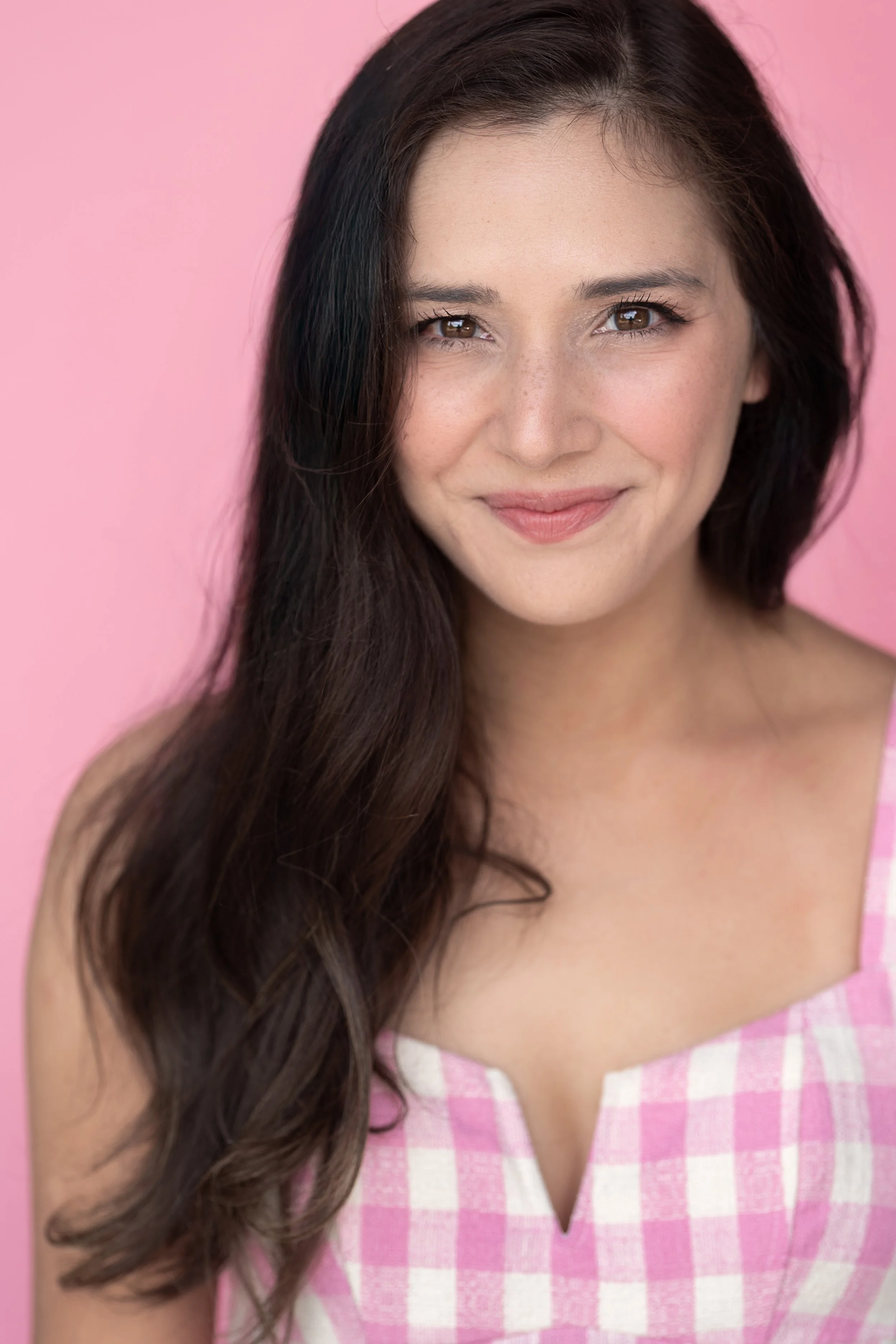 Close-up portrait of a young woman with long dark hair, smiling, wearing a pink and white checkered dress, against a pink background.