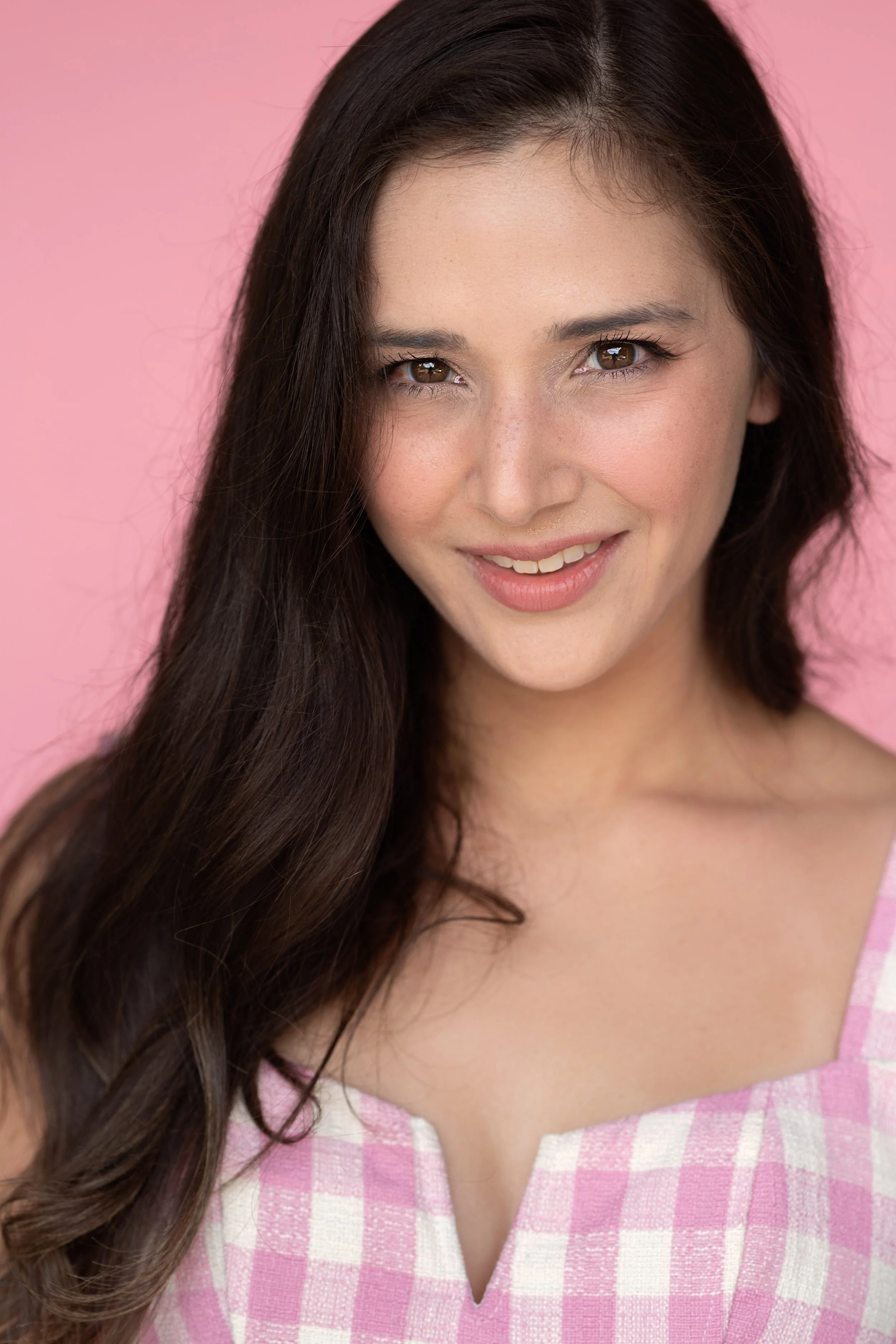 A woman with long brown hair, smiling, wearing a pink and white checkered top against a pink background.