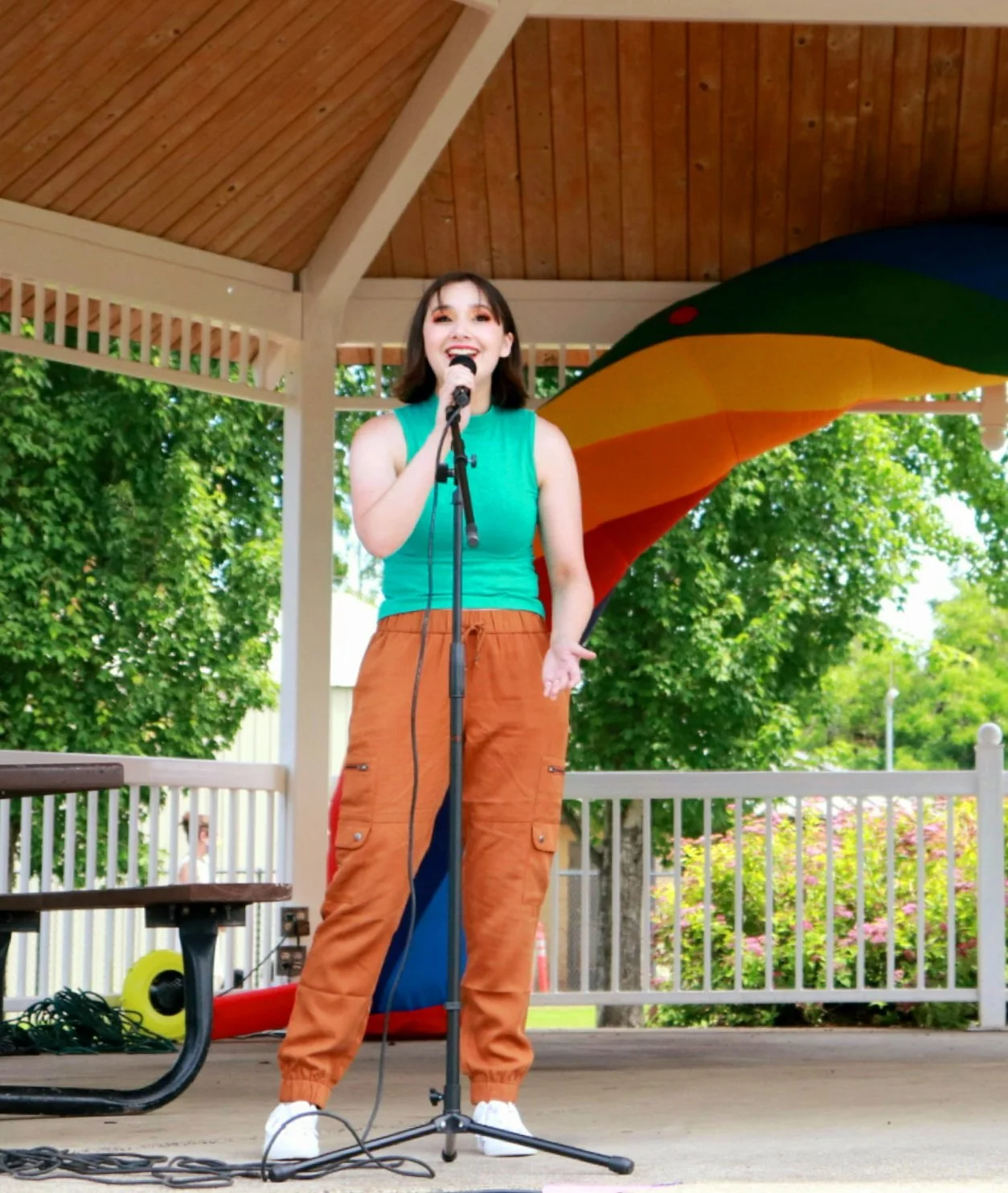Three people stand together in a colorful setting with flowers and stained glass, smiling and talking. The woman in the center wears a striped tank top and denim shorts, flanked by two men wearing hats.