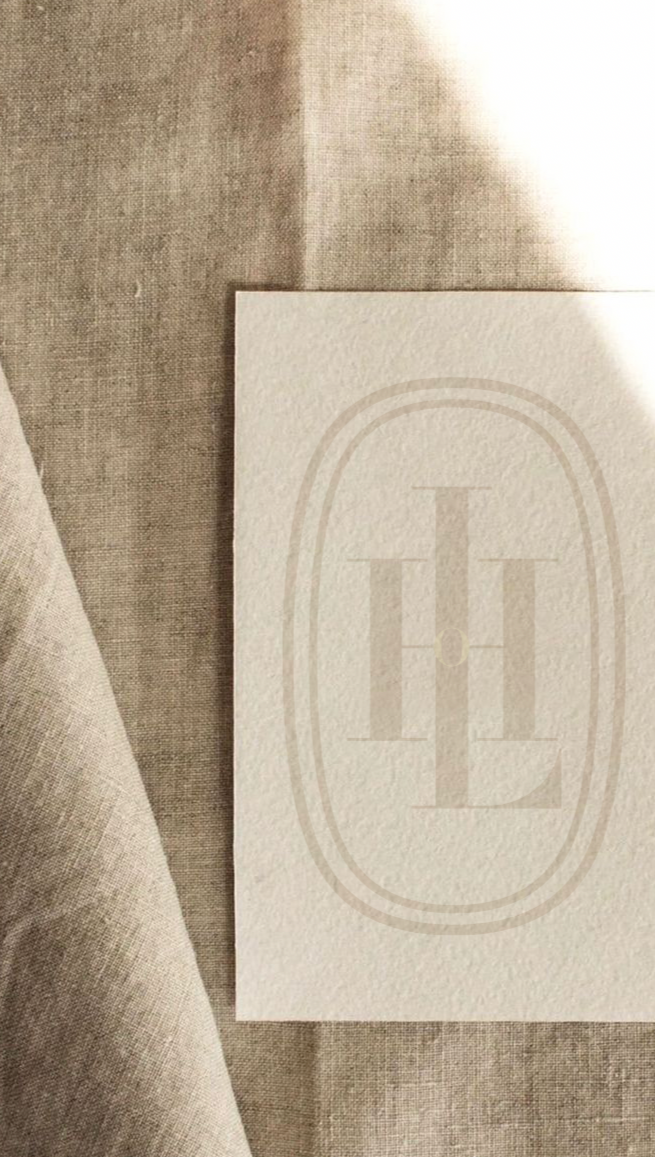 Close-up of a cream-colored textured paper with a faint, elegant monogram printed in the center. The paper is placed on a soft, beige fabric surface.