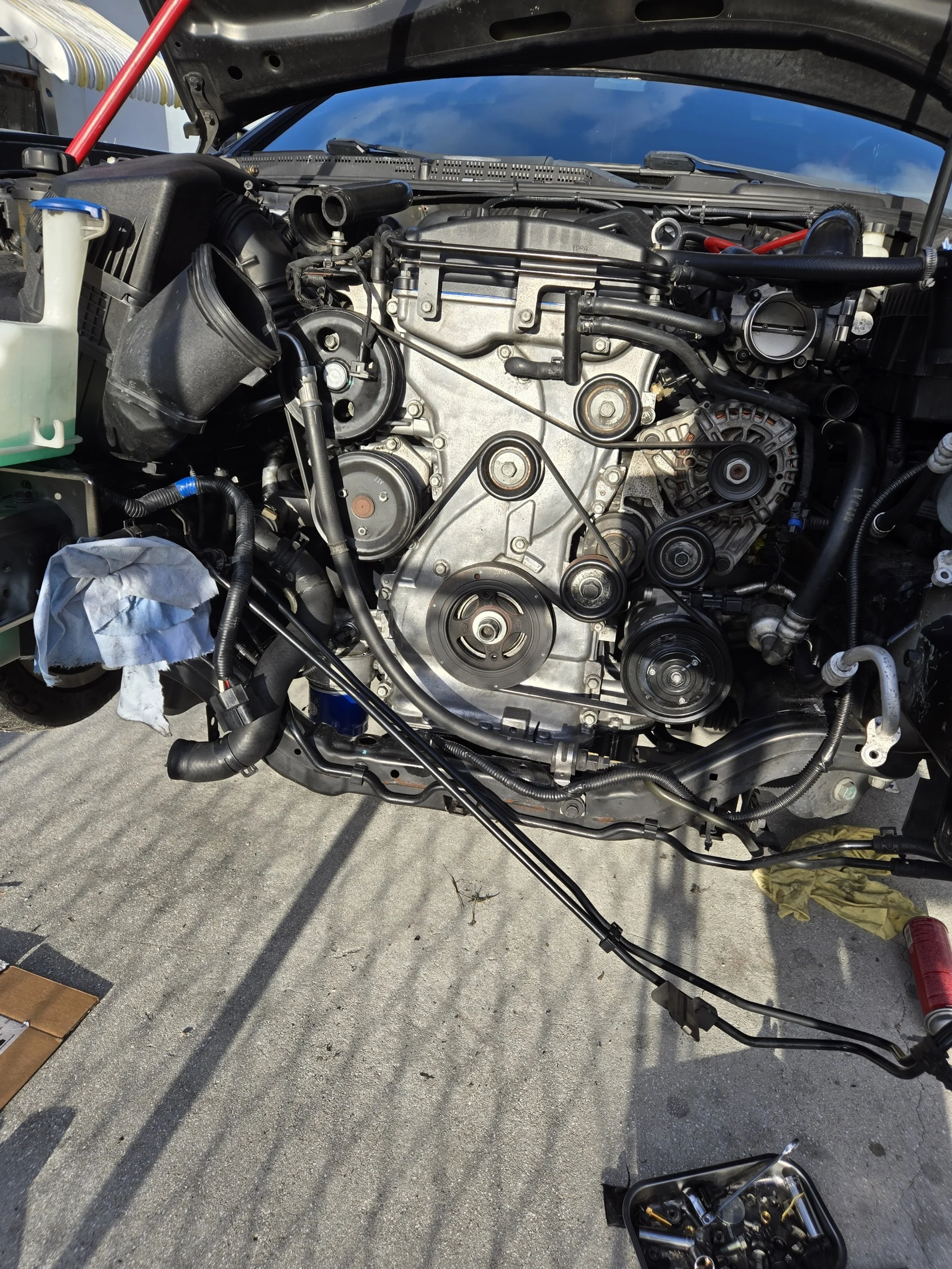 A car engine with the hood open, showing various components, belts, and pulleys, on a concrete surface with tools nearby.