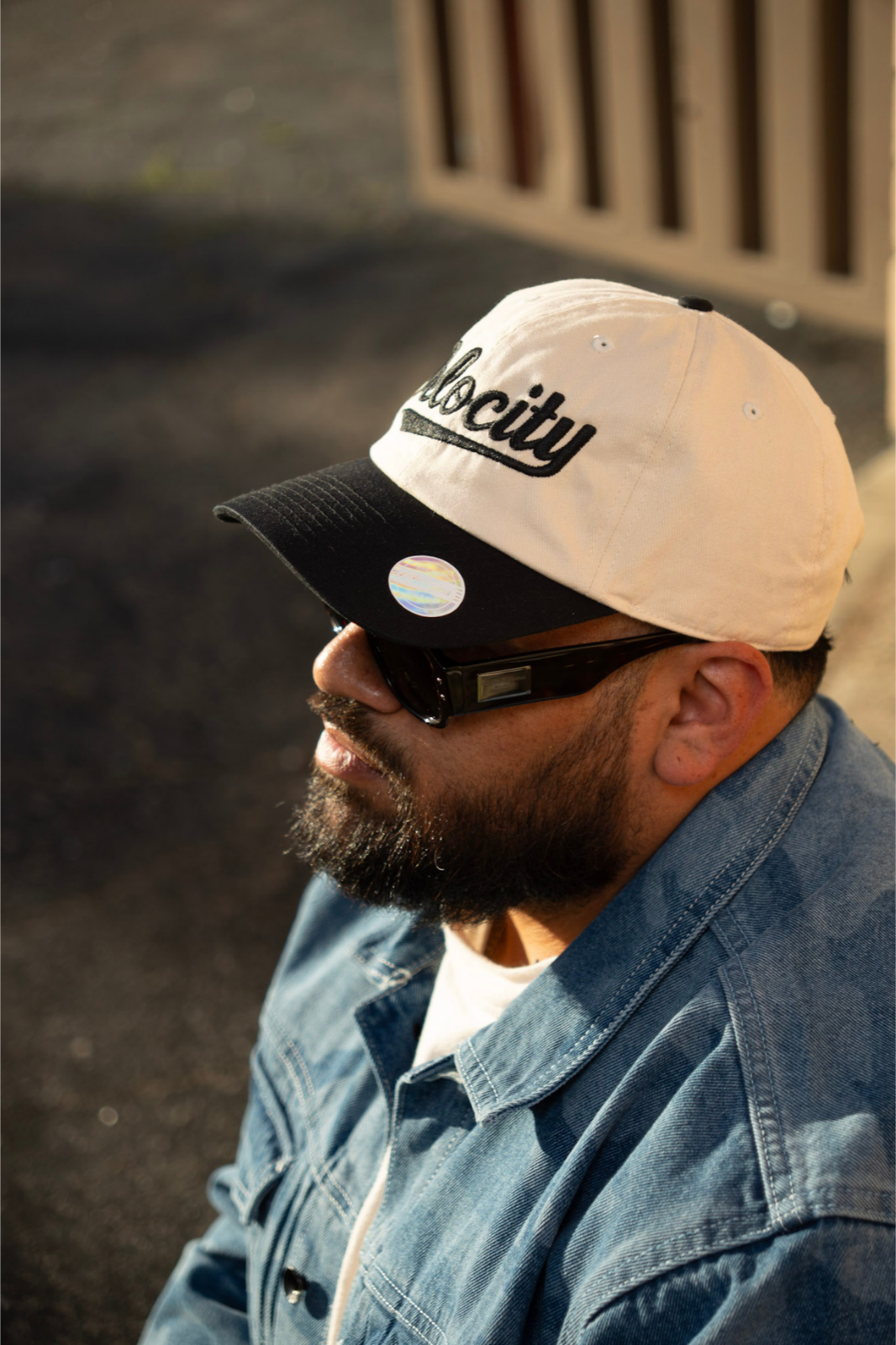 A man with a beard wearing sunglasses, a denim jacket, and a baseball cap with the word 'mobcity' on it, sitting outdoors during daytime.